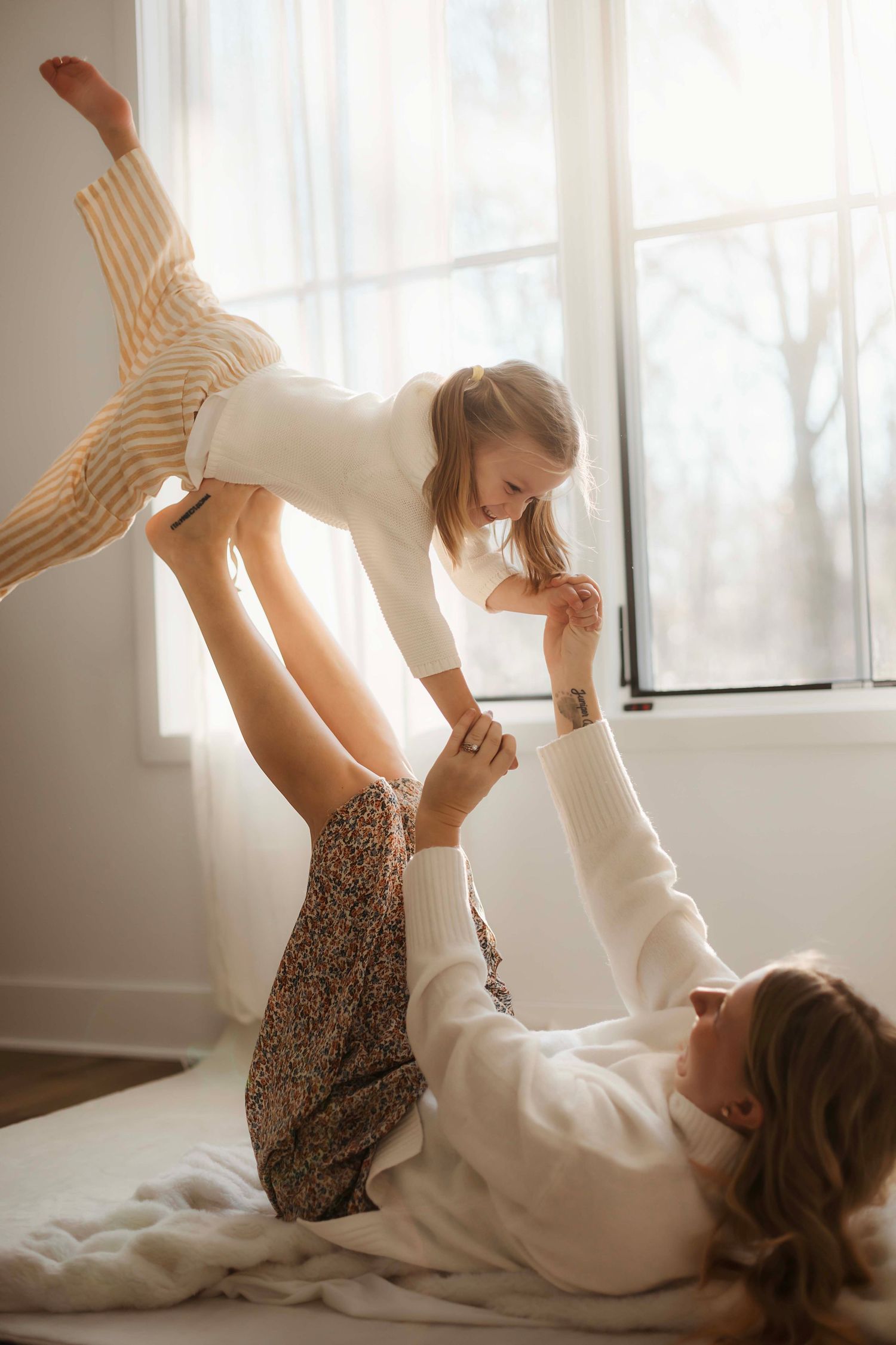 A playful moment captured in soft light as two people balance and play together on a bed by a window.