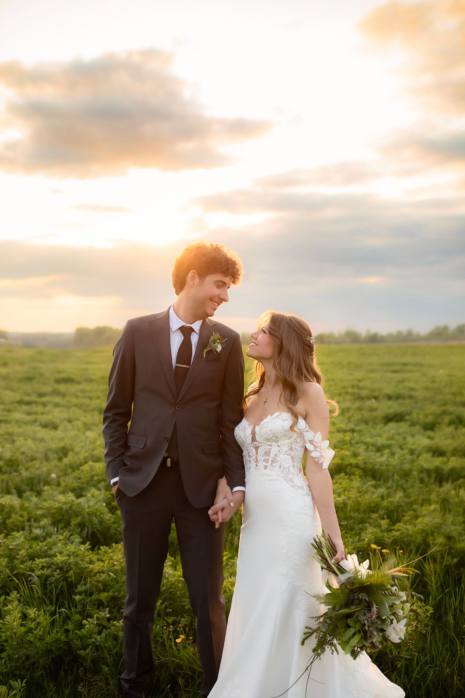 A bride and groom hold hands in a green field at golden sunset, sharing a loving gaze in elegant wedding attire.