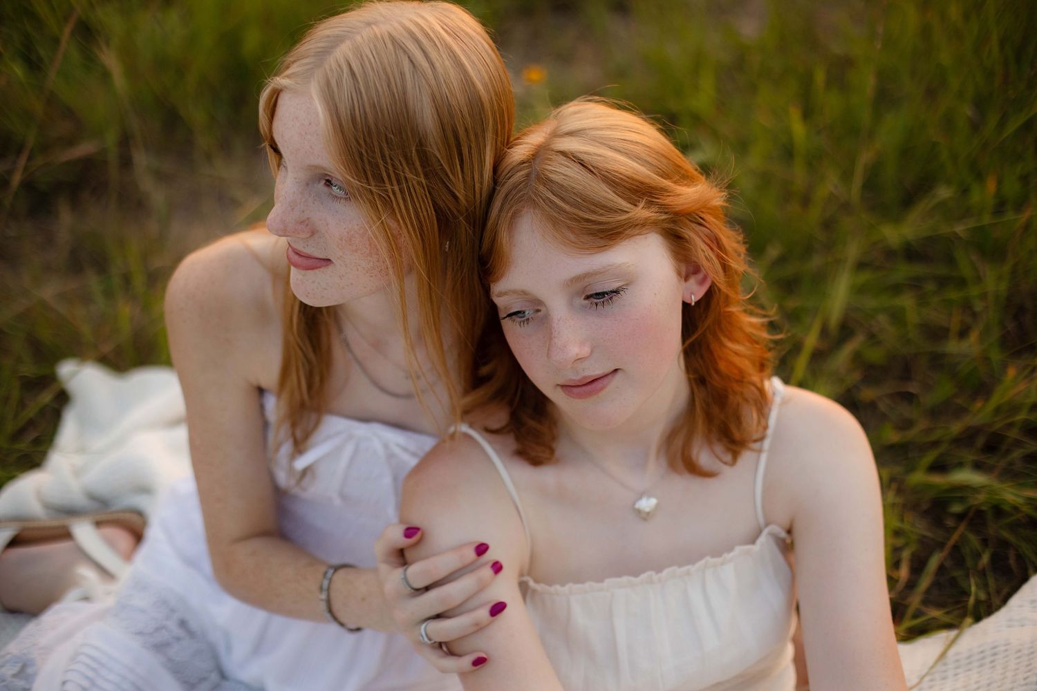 Two red-haired girls in white dresses sit together on a blanket in a grassy field during golden hour.