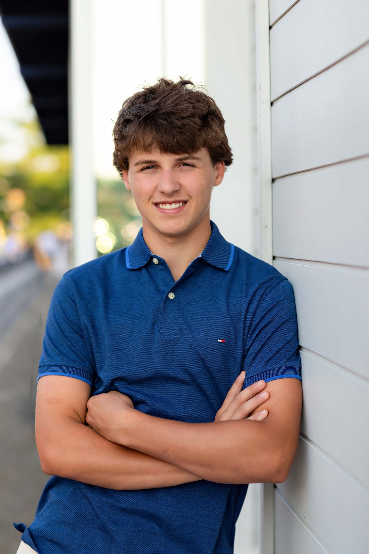 Smiling teen boy in blue Tommy Hilfiger polo with arms crossed leaning on wall.