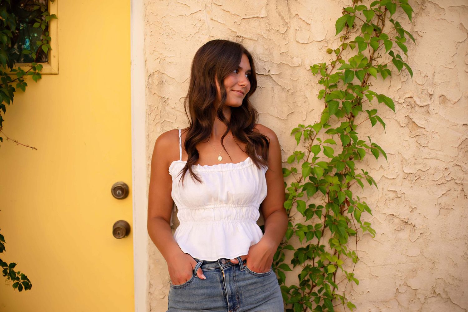 Young woman in white crop top and jeans smiling outdoors near yellow door with green ivy wall.