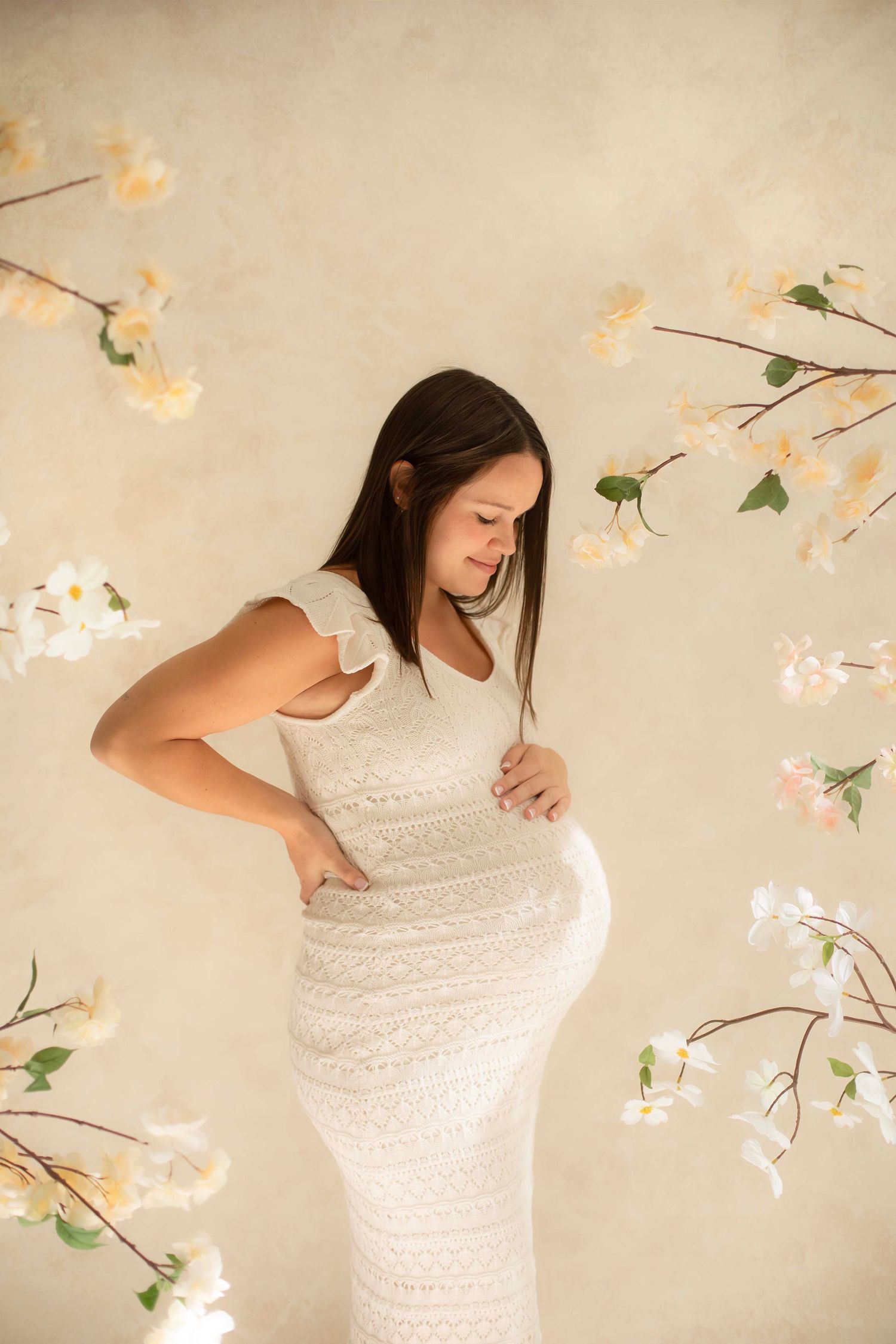 Pregnant woman in white lace dress cradling baby bump against floral backdrop.