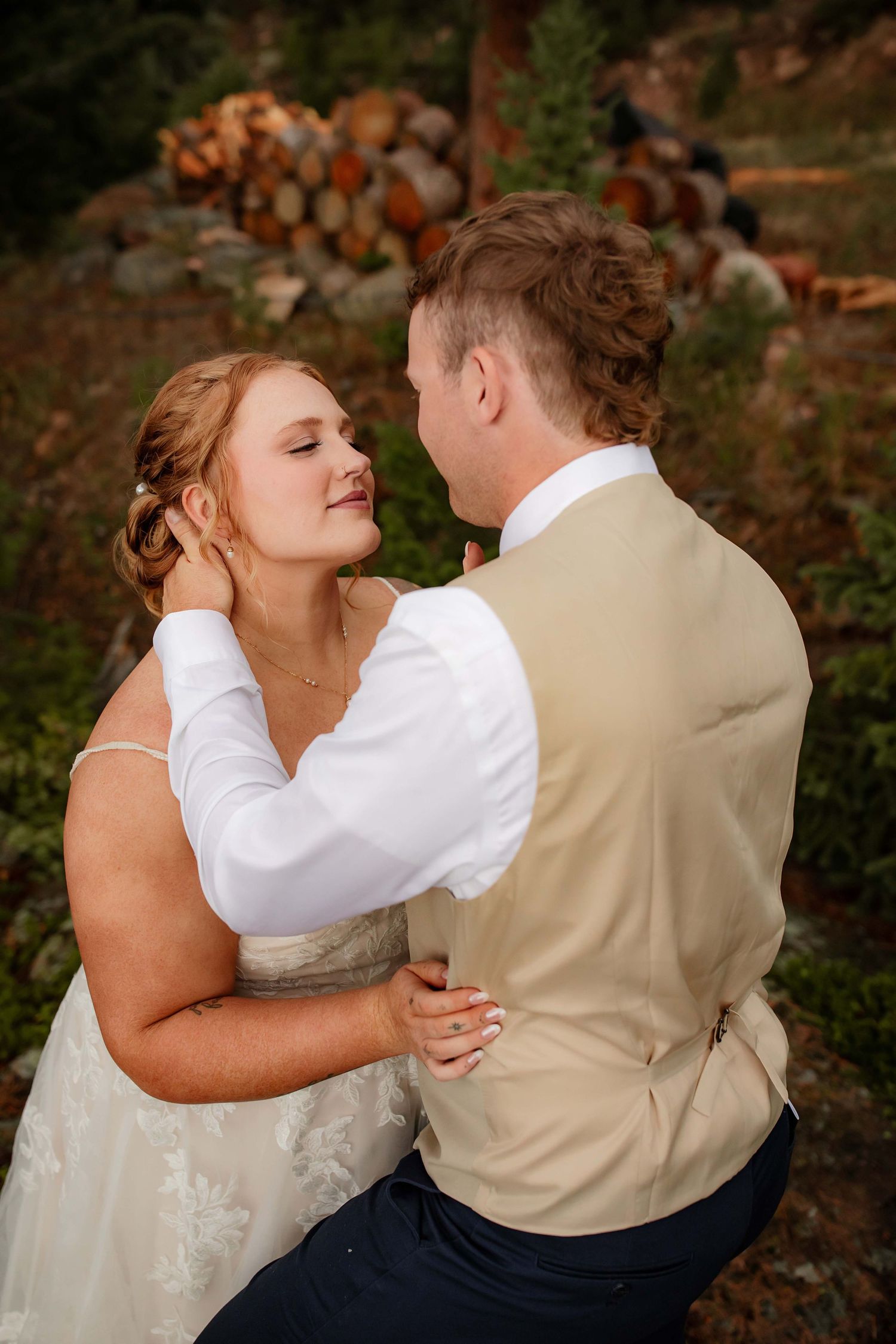 A romantic moment between newlyweds embracing outdoors with stacked firewood in the background.