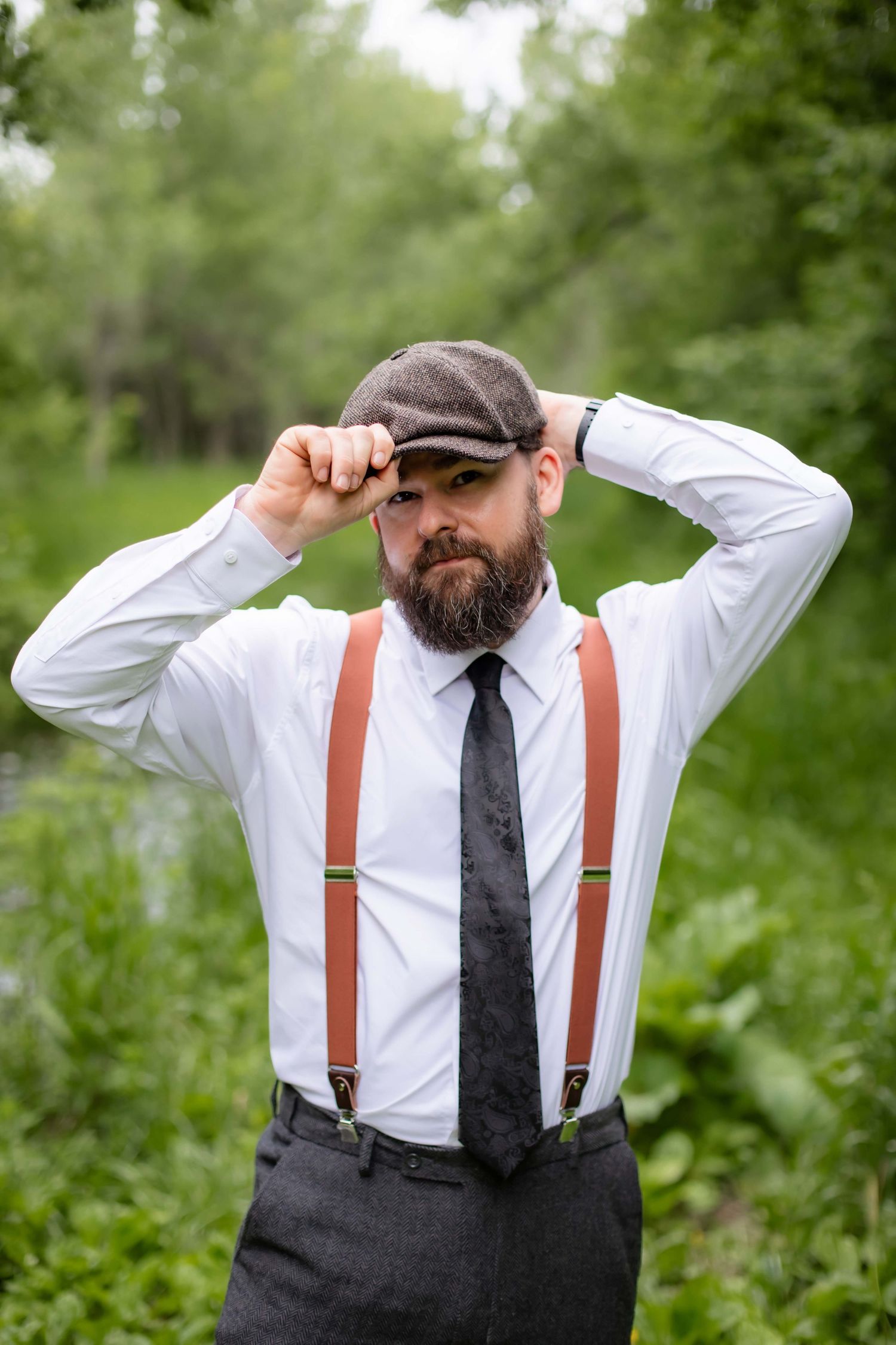 A groom in suspenders and flat cap adjusts his hat while wearing a white dress shirt and tie.