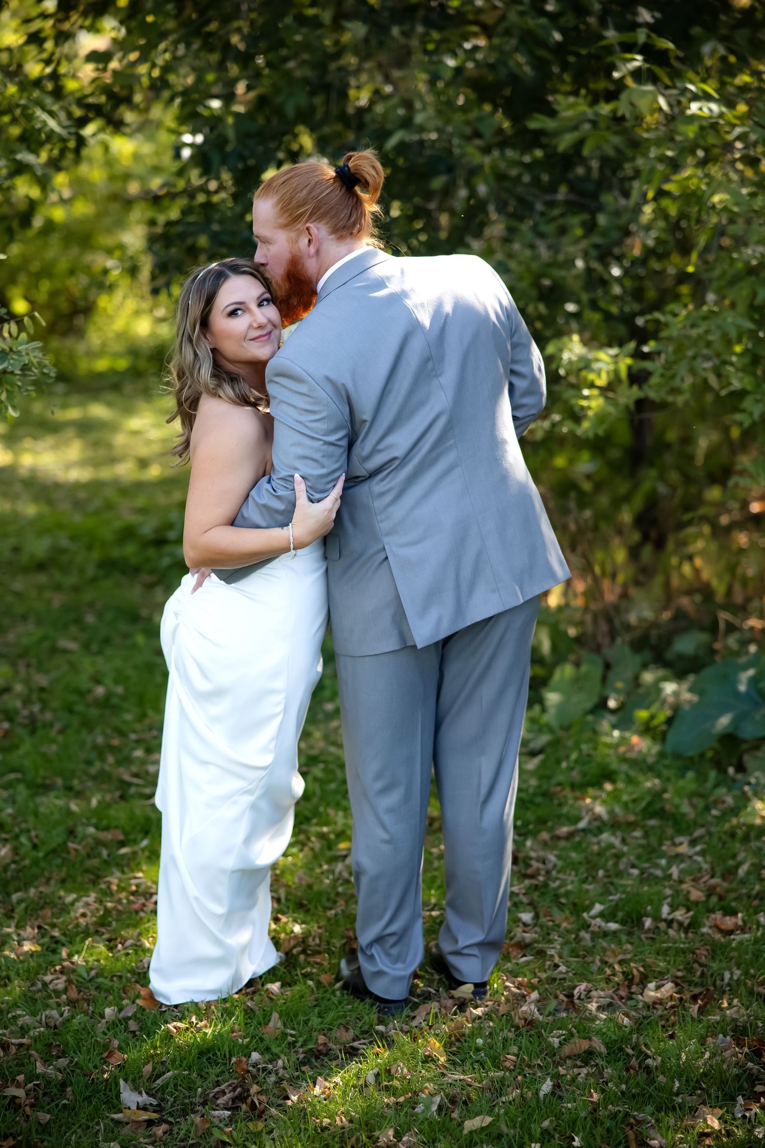 A couple shares a playful moment in a garden setting wearing white and gray formal attire.