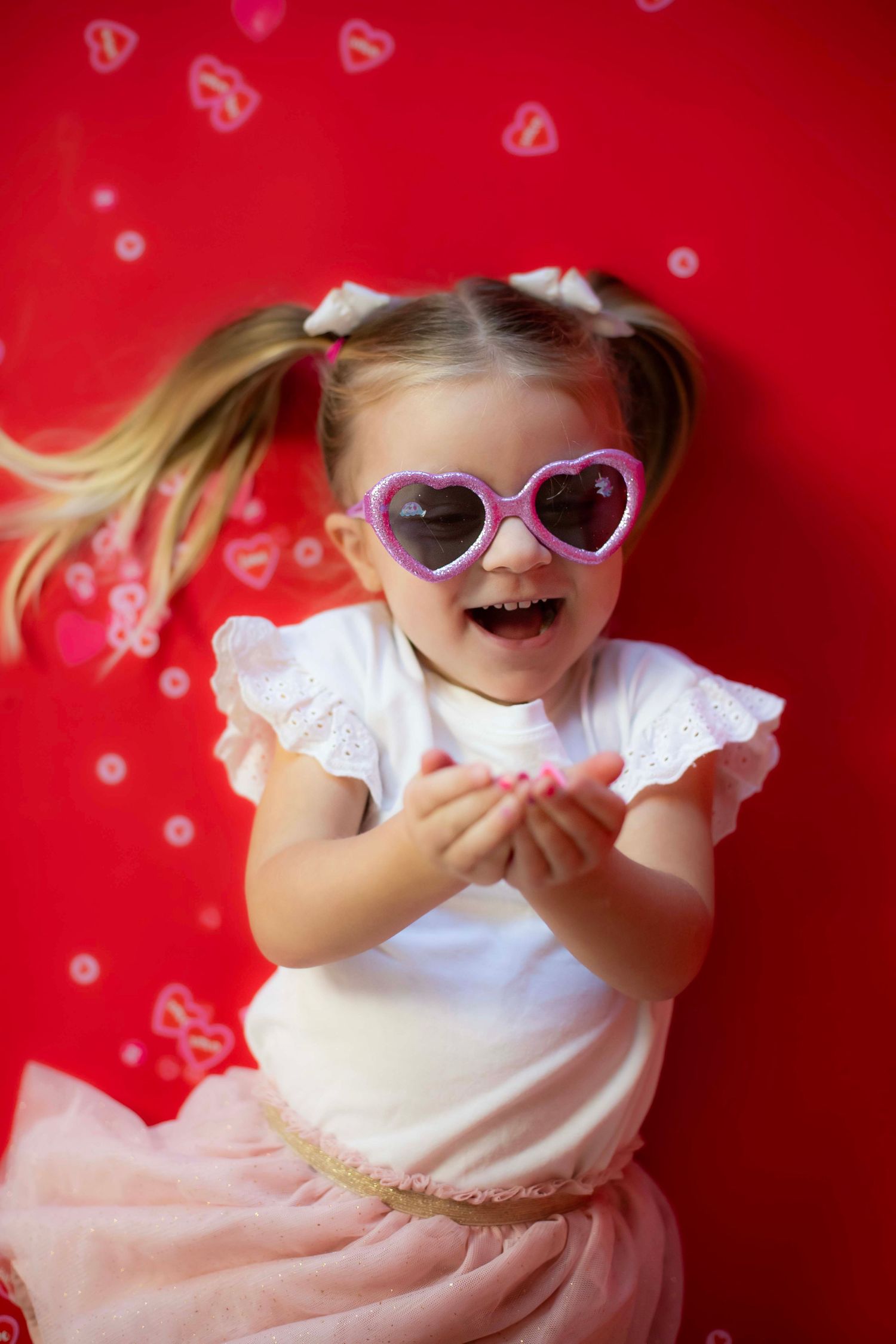 Excited pose with heart sunglasses against red backdrop with floating heart bokeh effects.
