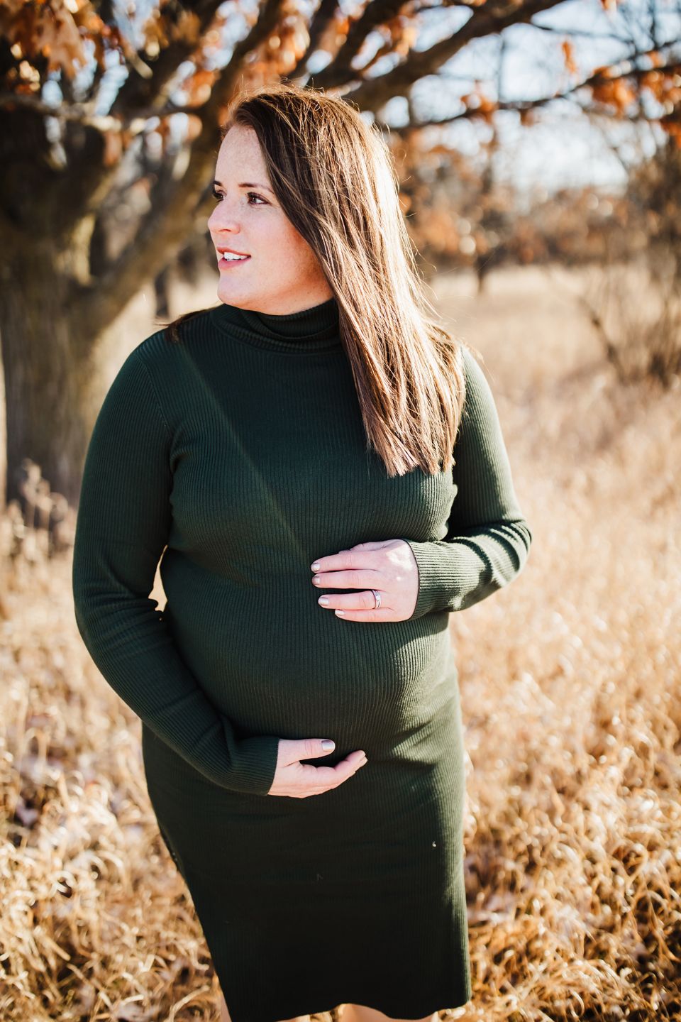 Woman in dark green sweater dress poses in autumn field with golden grasses and tree branches in background.