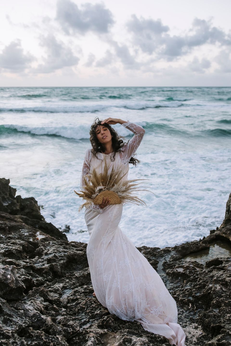 A bride in flowing white dress poses with pampas grass against dramatic ocean waves and rocky coastline.