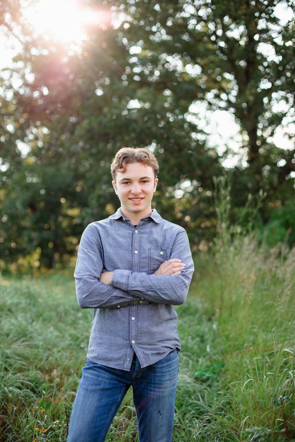 Person in blue button-down shirt poses with crossed arms in field at sunset.