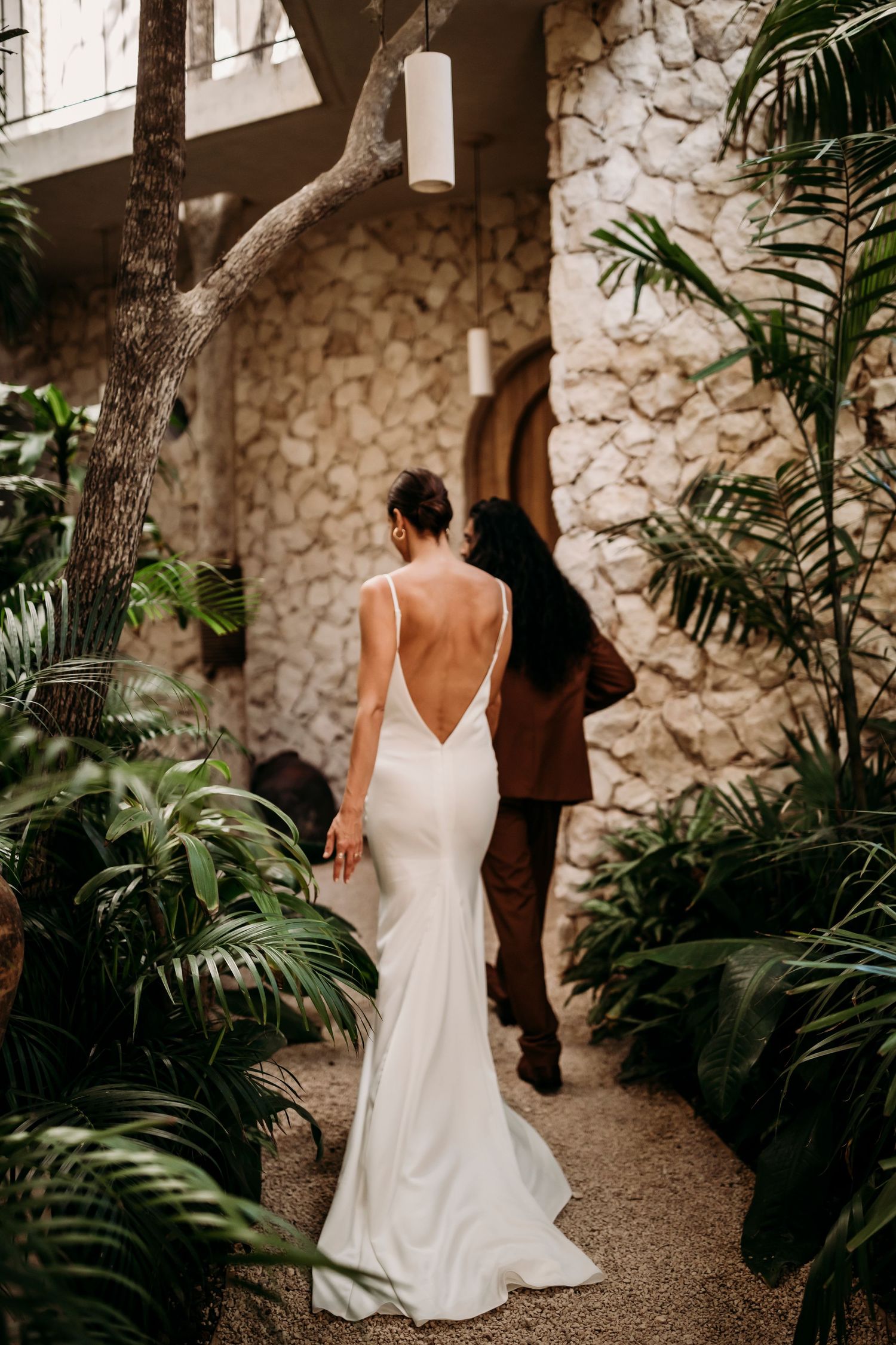 Woman in elegant white wedding dress walking through tropical garden path with stone walls.
