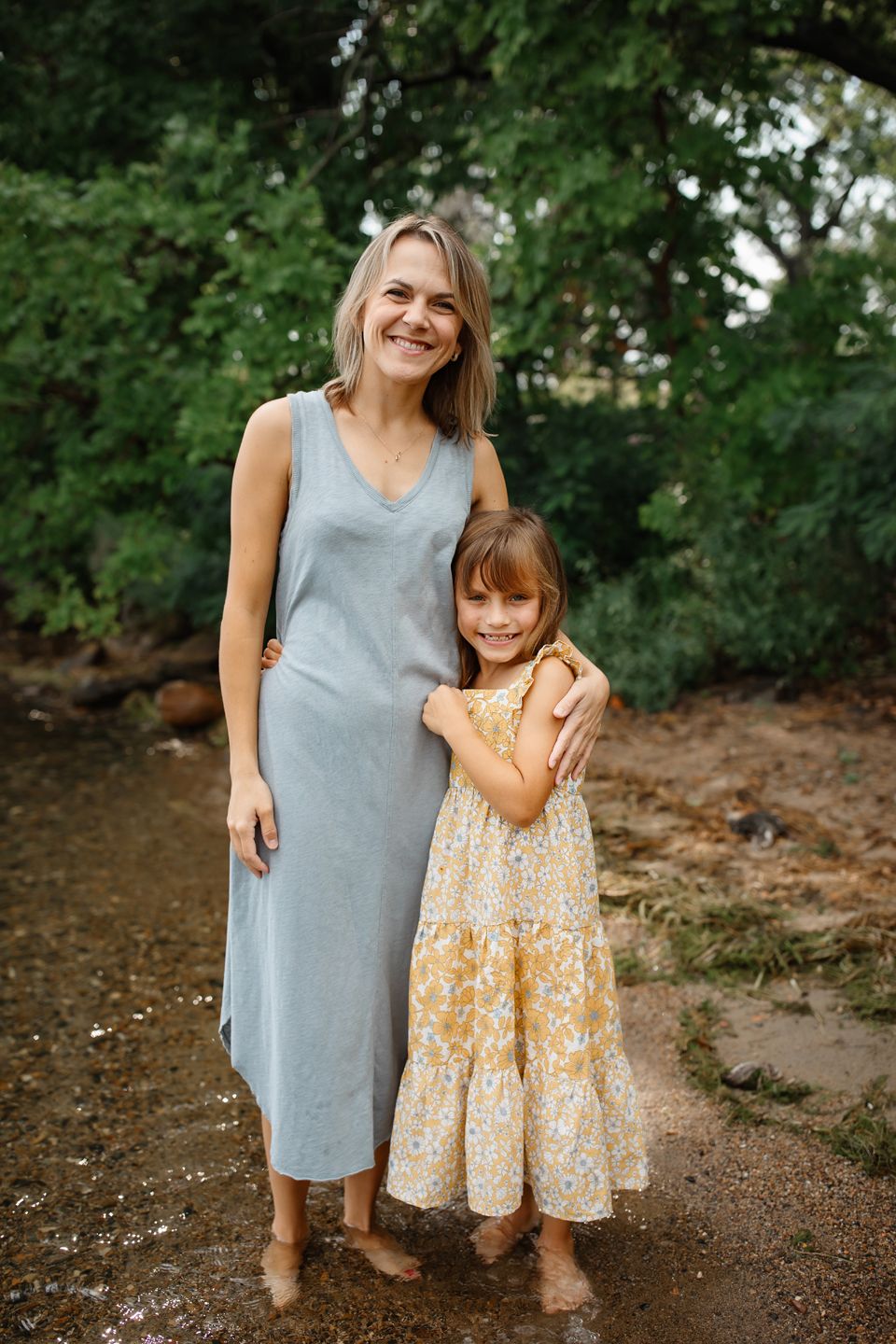 A woman in a gray maxi dress and a child in a floral yellow dress pose together on a dirt path surrounded by greenery.