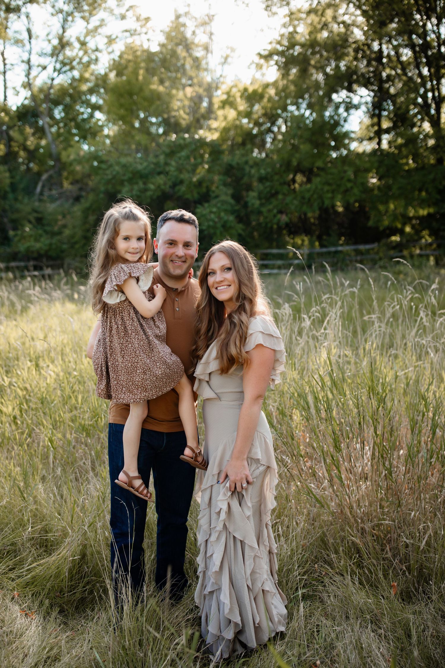 A family poses together in a field of tall grass at sunset with trees in the background.
