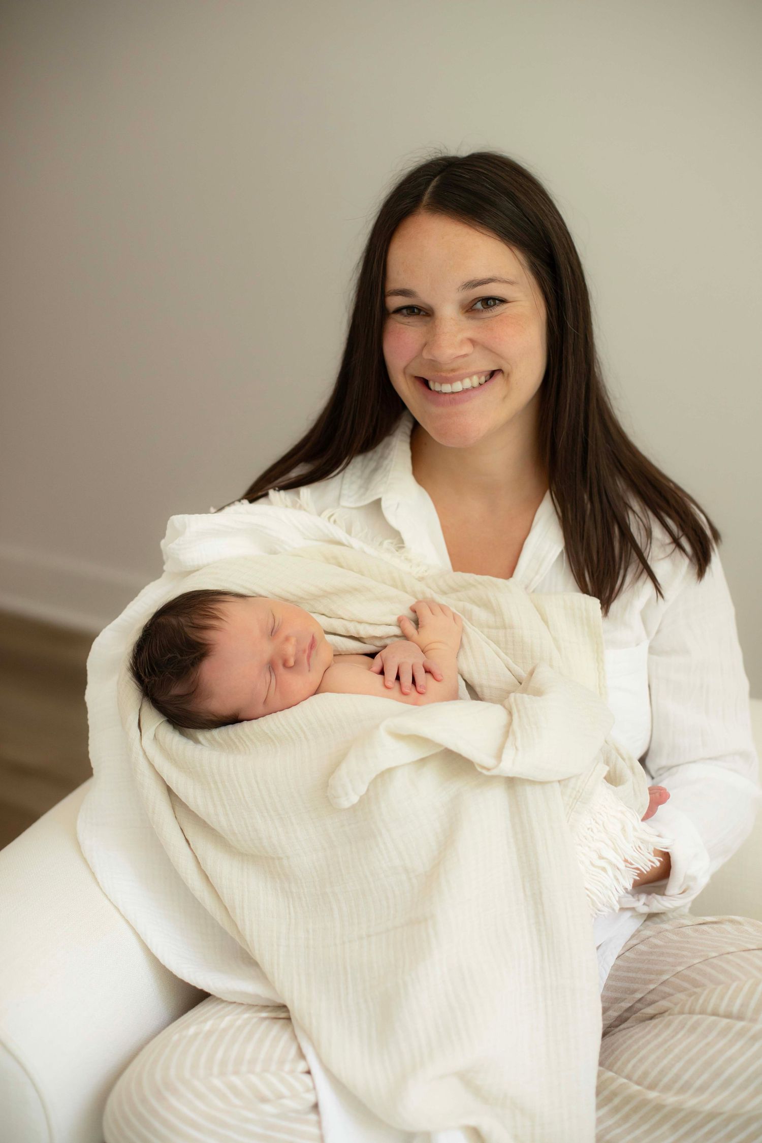 Smiling mother with long dark hair holds sleeping newborn baby wrapped in cream blanket.