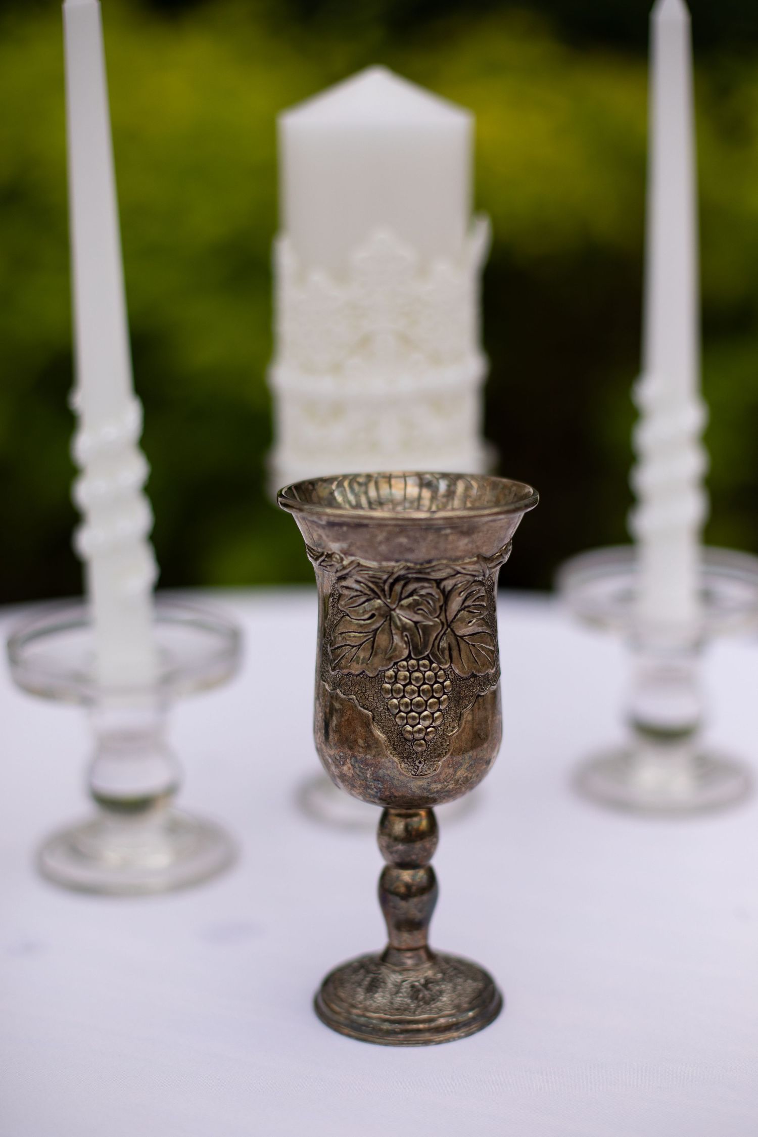 Ornate antique metal chalices and candlesticks arranged in a row with white candles against a blurred green background.