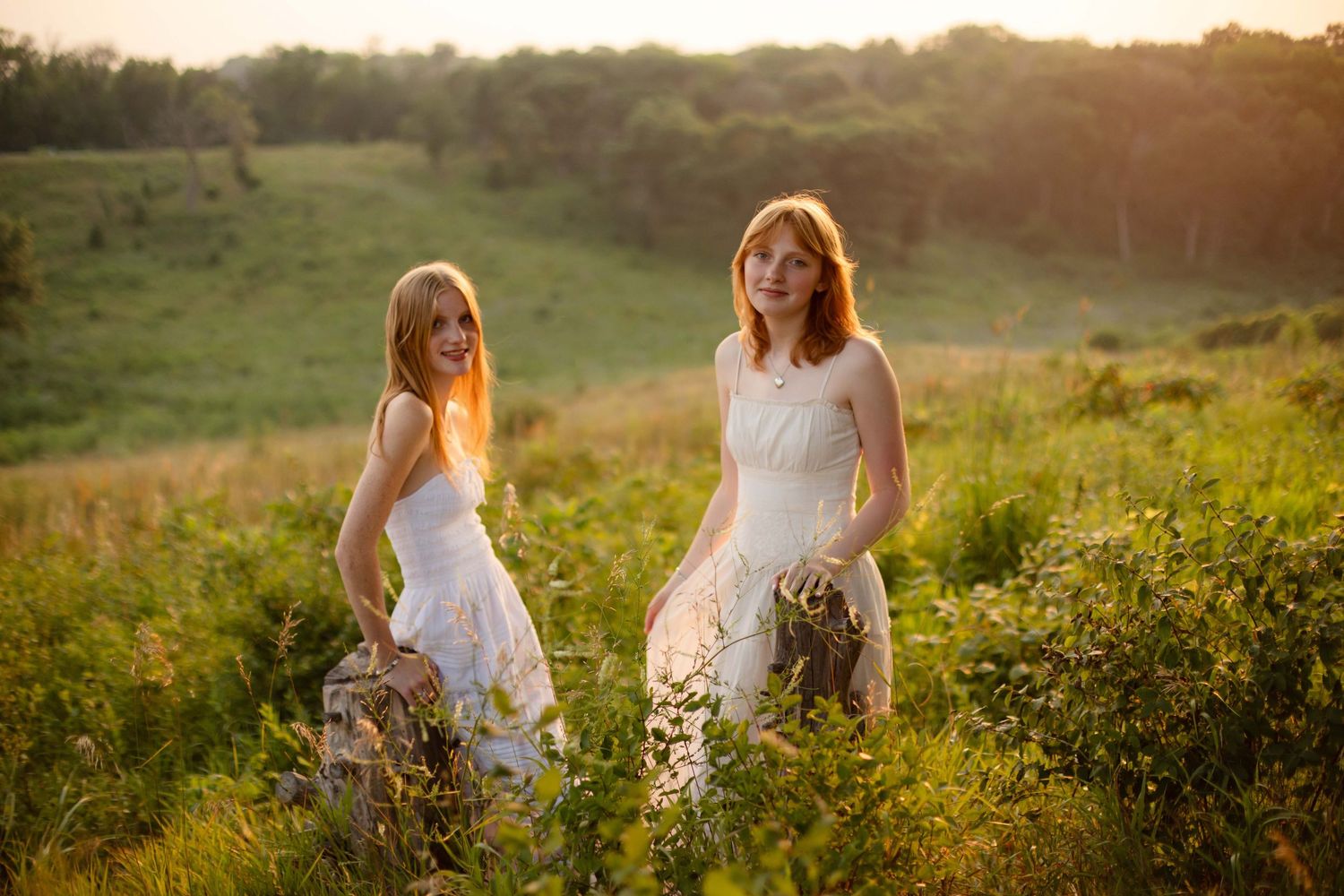 Two young women in white dresses standing in a sunlit green field during golden hour.
