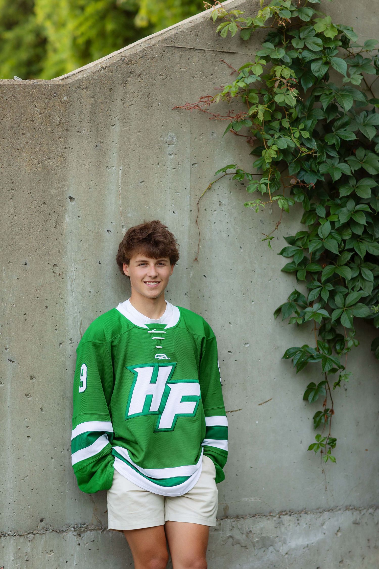 Young athlete in green HF hockey jersey number 9 posing against concrete wall with ivy outdoors.