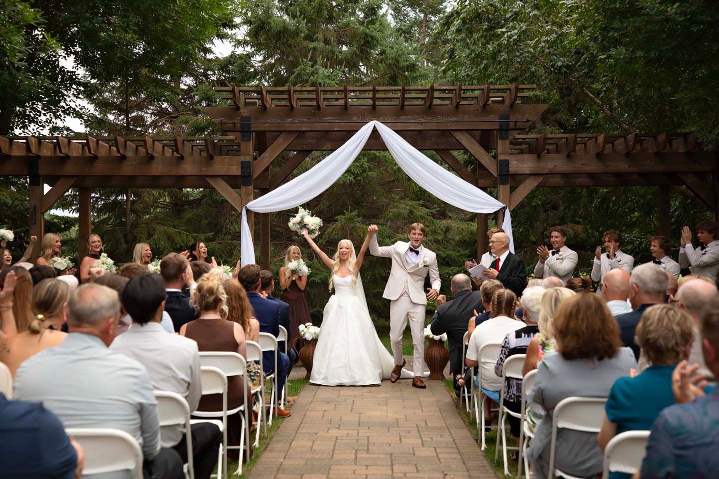 Bride and groom raise hands in celebration at outdoor garden wedding ceremony under wooden pergola with white draping.