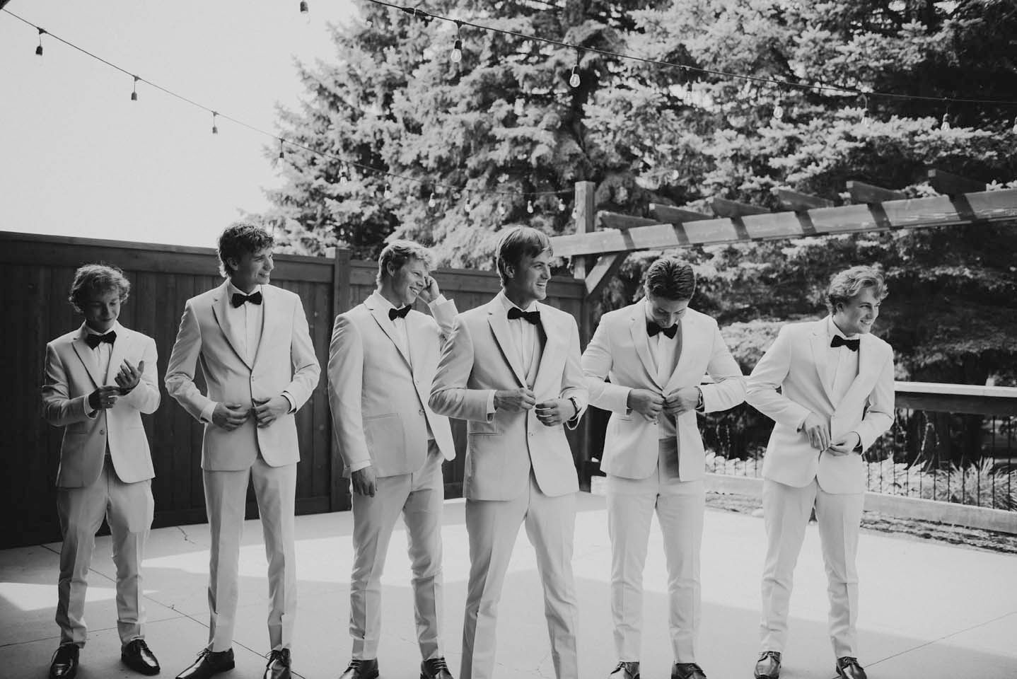 Six groomsmen in white suits and bow ties pose in a line outdoors in a black and white wedding photo.