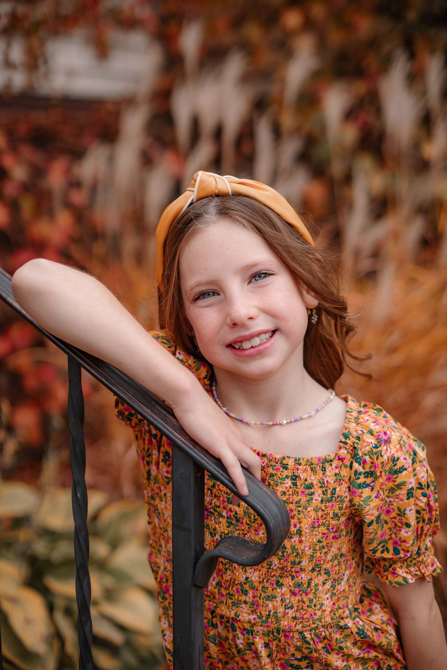 Portrait in floral dress against autumn foliage with warm lighting and iron railing.
