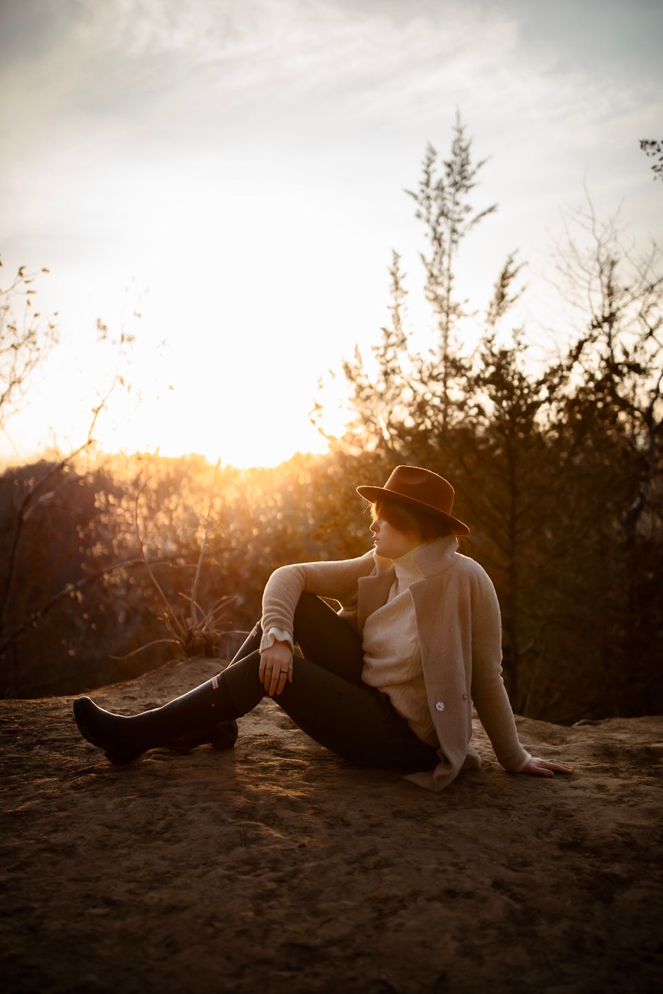 A person in a hat and neutral clothing reclines on rocky ground during golden hour.