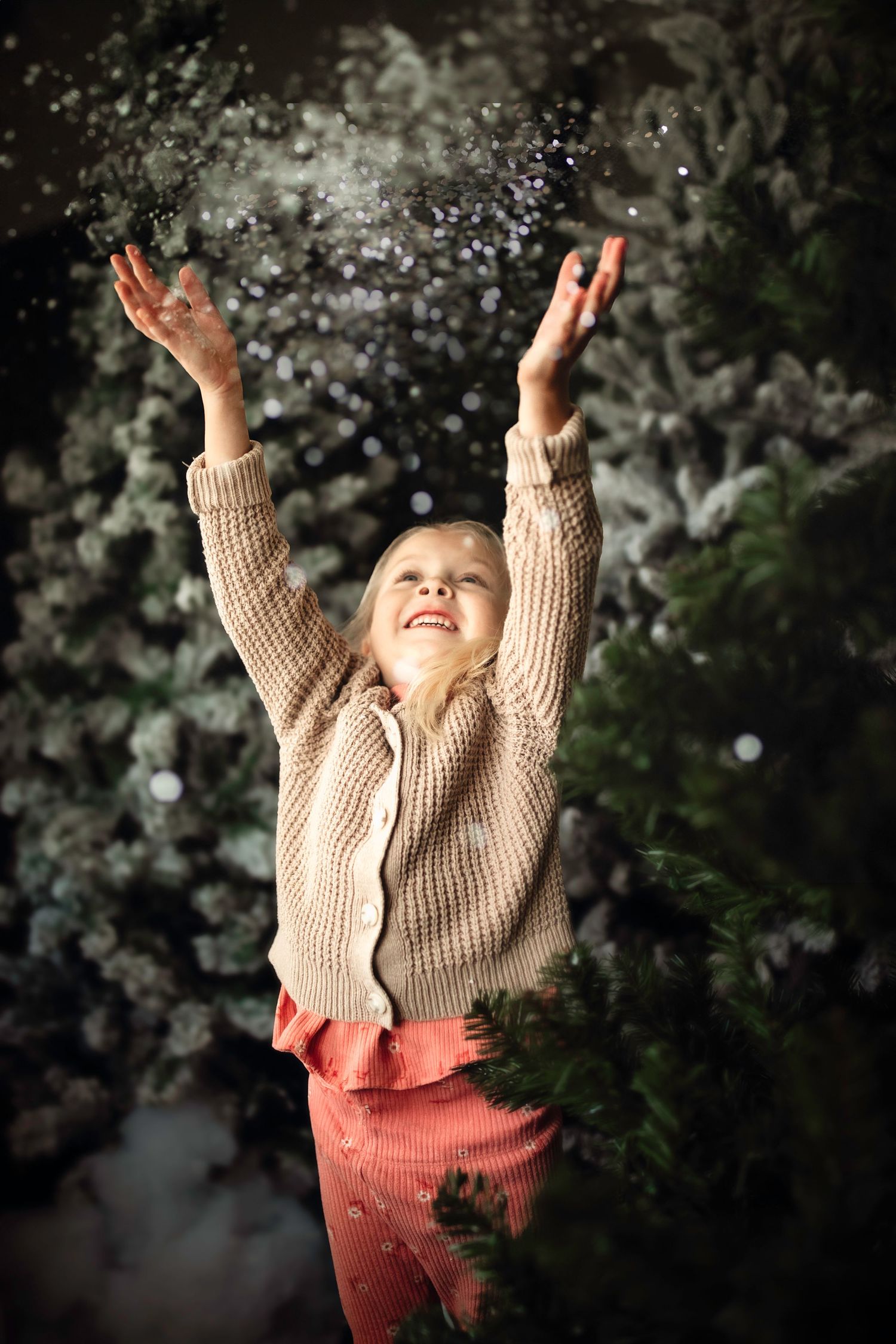 Child in beige sweater throwing snow in the air with joy against dark winter background.