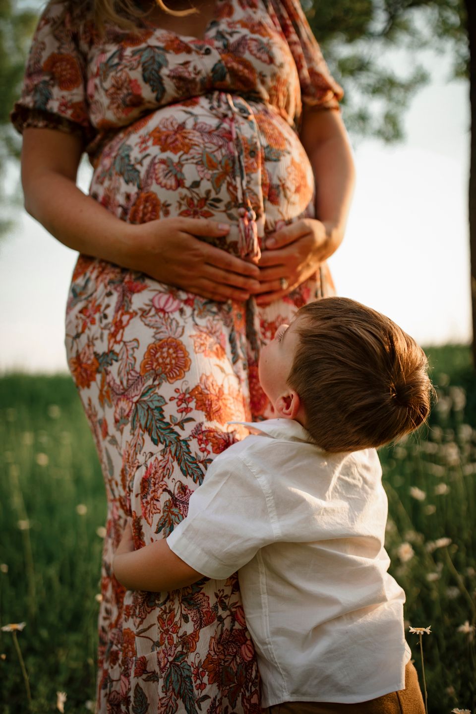 Child in white shirt hugging and embracing pregnant mother wearing floral dress in sunlit meadow at sunset.