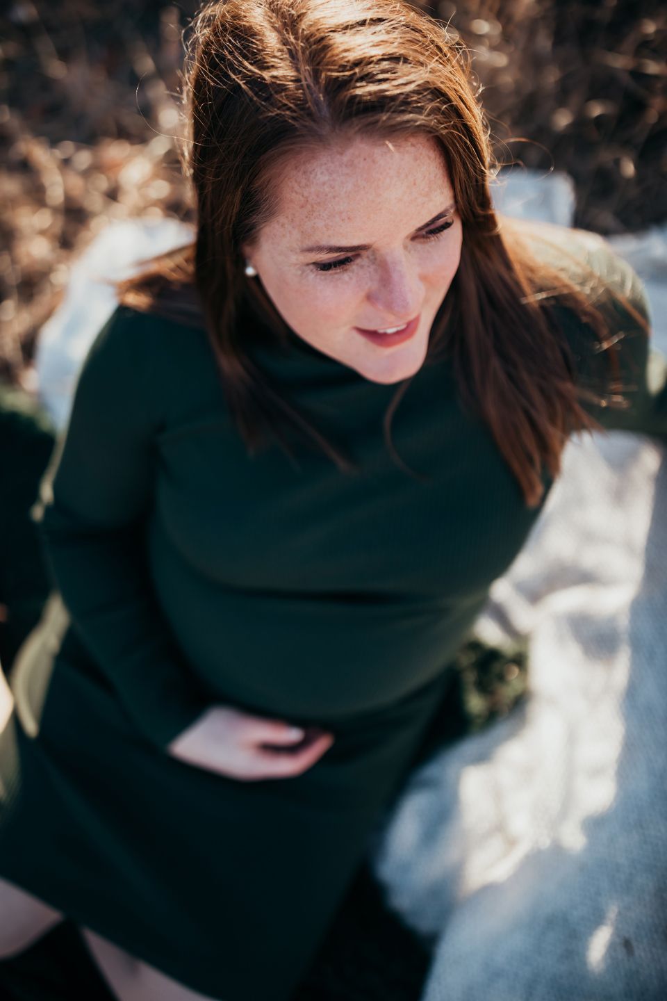 Woman in dark green sweater sitting outdoors in natural lighting with dappled sunlight falling across the scene.