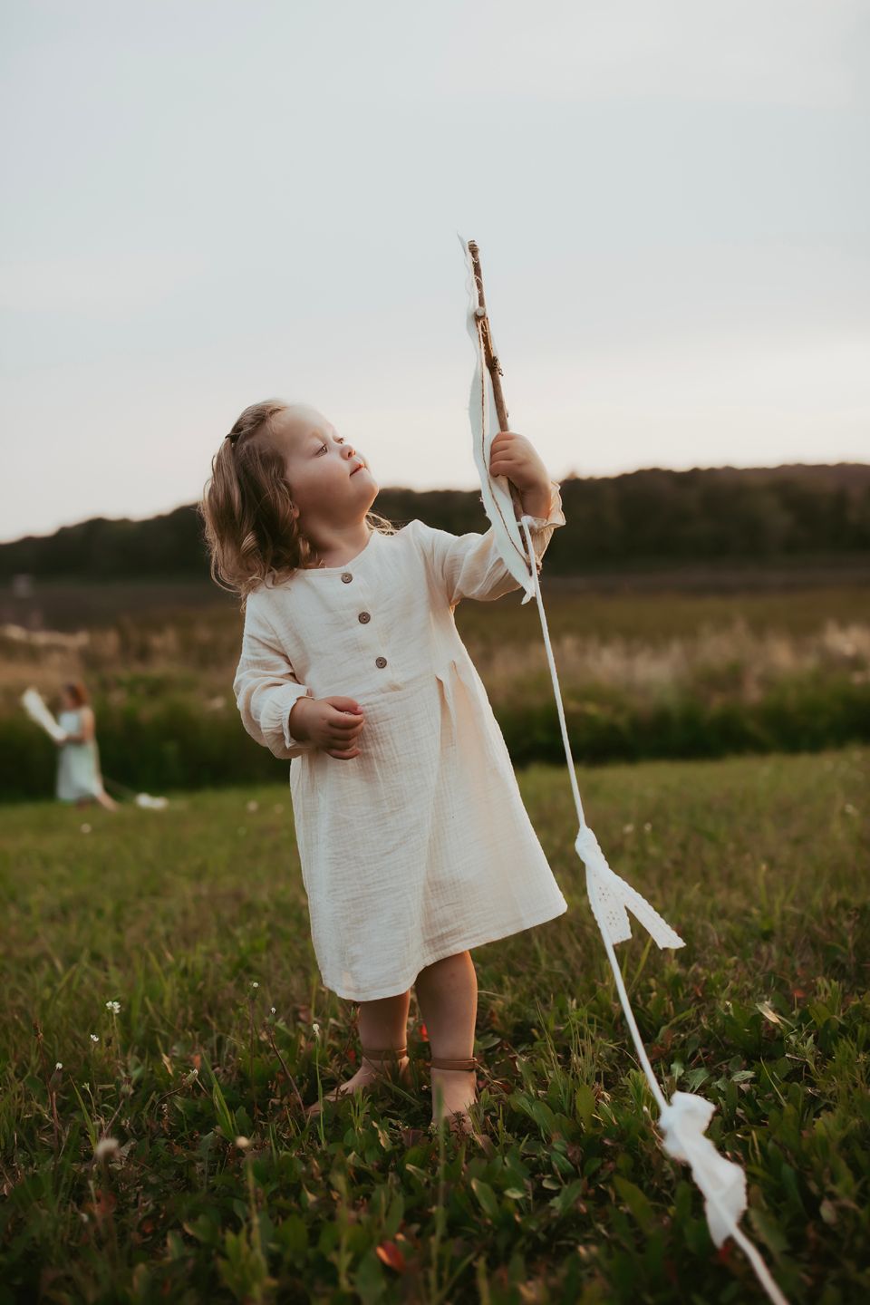 A small figure in a white dress holds a decorative branch in a meadow at dusk.
