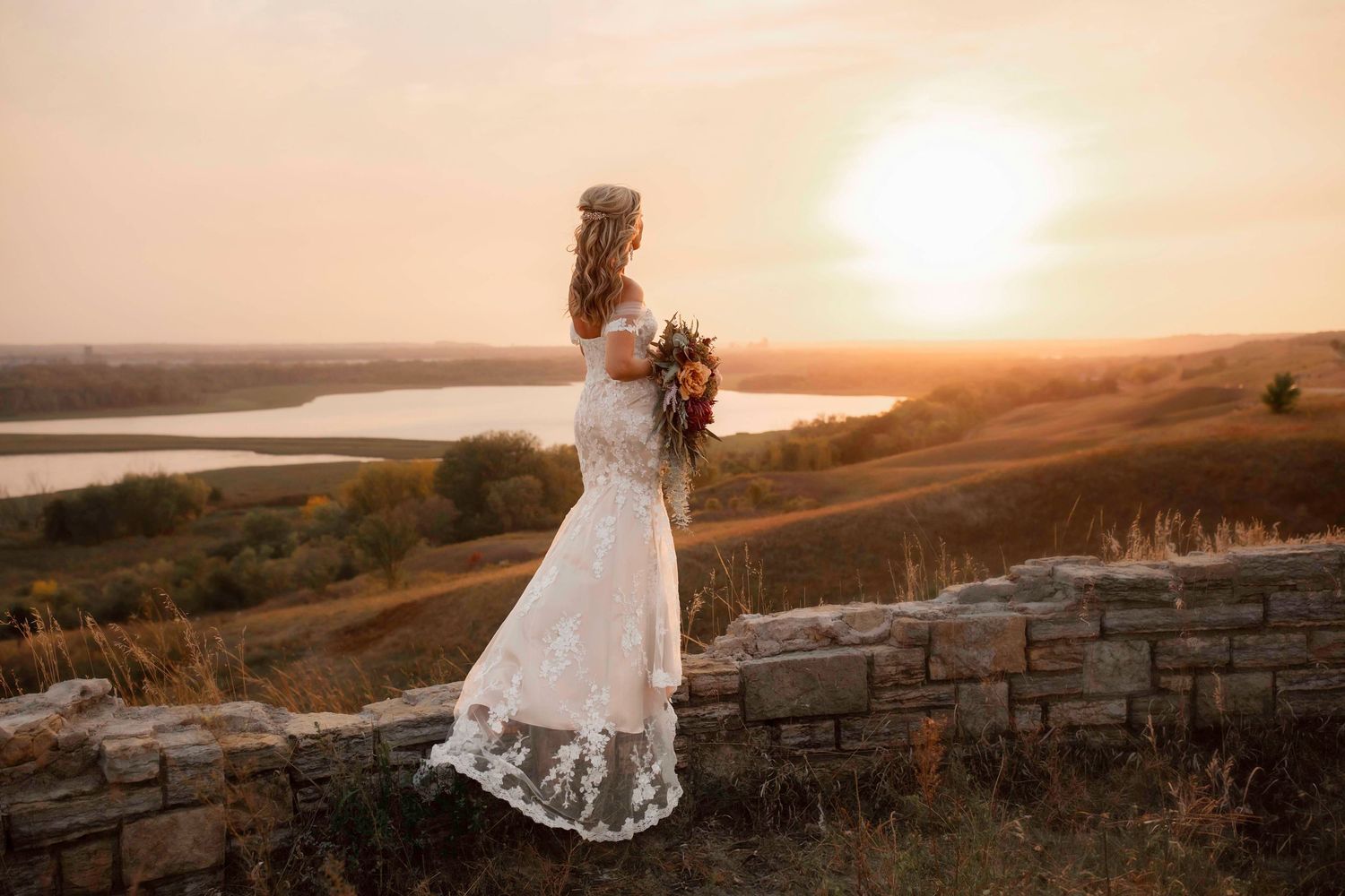 Bride in flowing lace gown stands on stone wall silhouetted against sunset and lake.