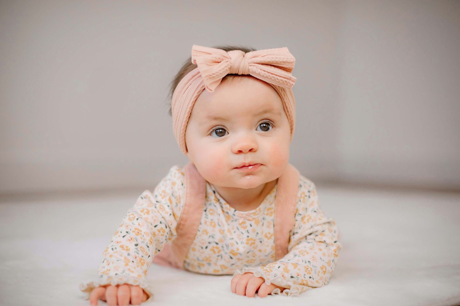 Baby wearing pink bow and floral outfit lying on gray surface.