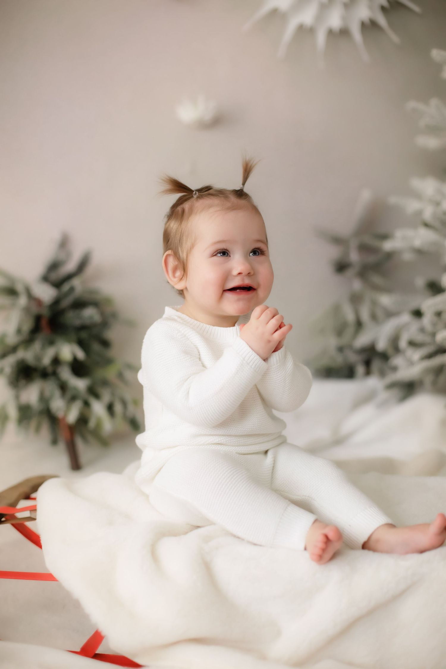 Baby in white outfit sits happily on white blanket with holiday decorations.