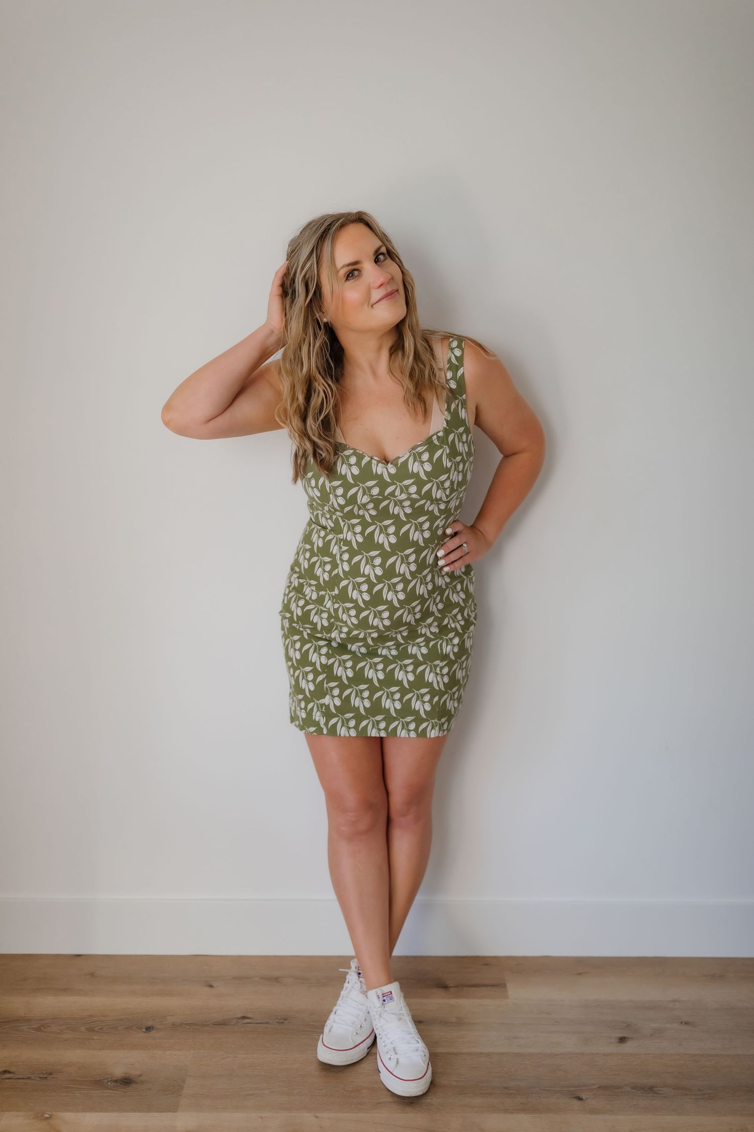 Casual pose in olive green patterned dress and white sneakers against neutral wall.