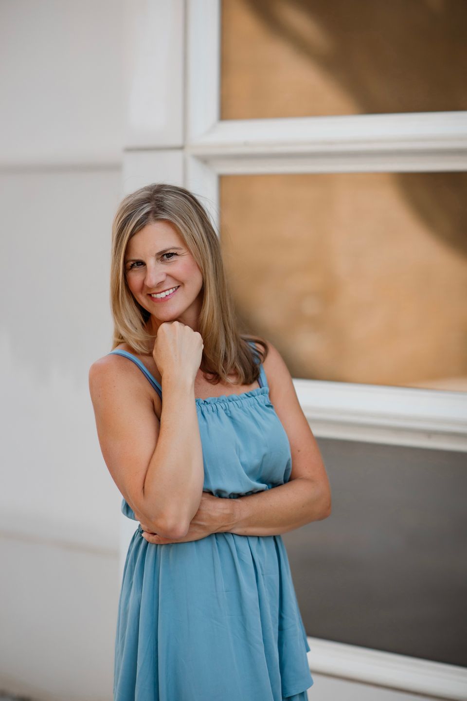 Portrait of someone wearing a blue summer dress posing against a white wall with natural lighting.