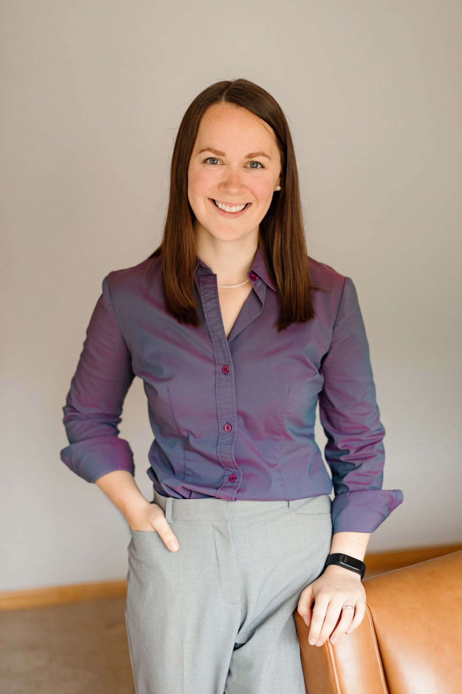 Business professional in purple shirt and gray slacks poses against warm interior background.