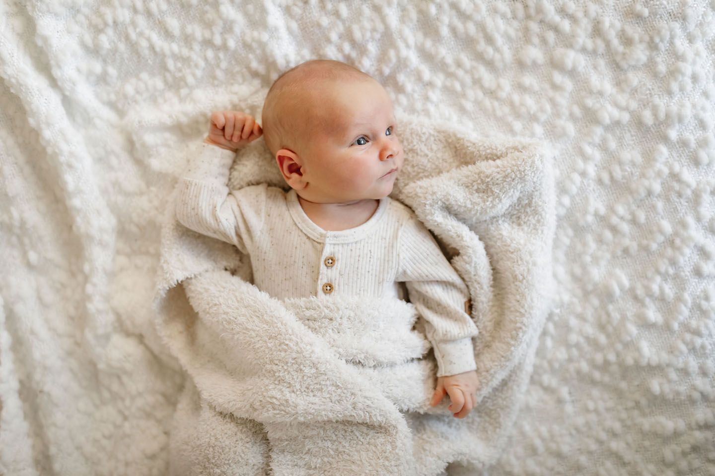 Baby in cream henley outfit lying on plush white bedding looking to the side.