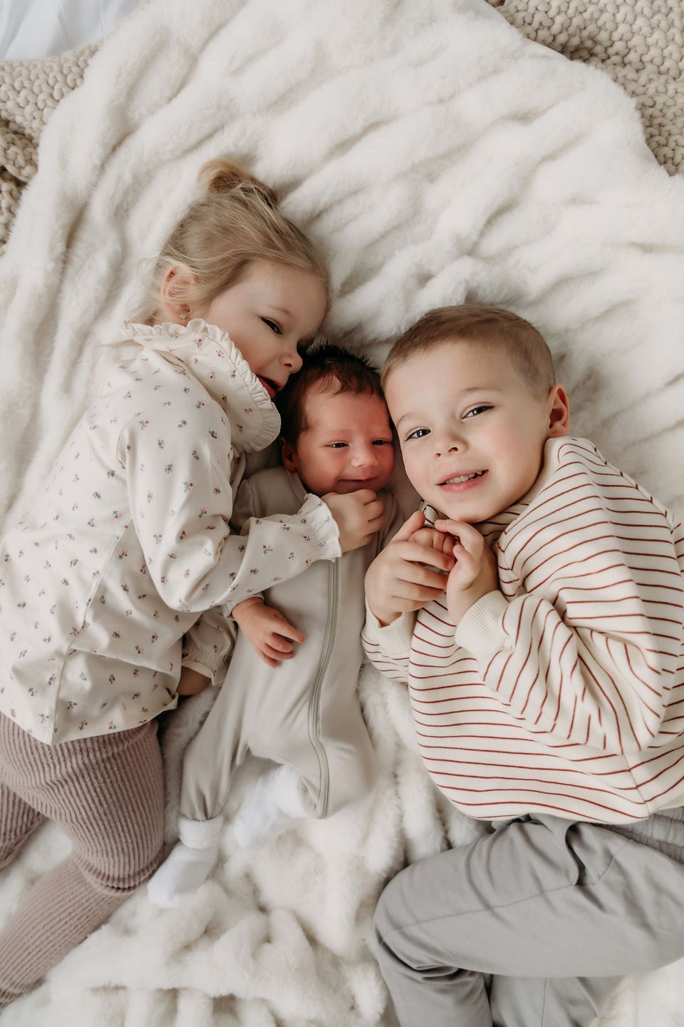 Three young children cuddle together on white bedding, dressed in cozy sleepwear.