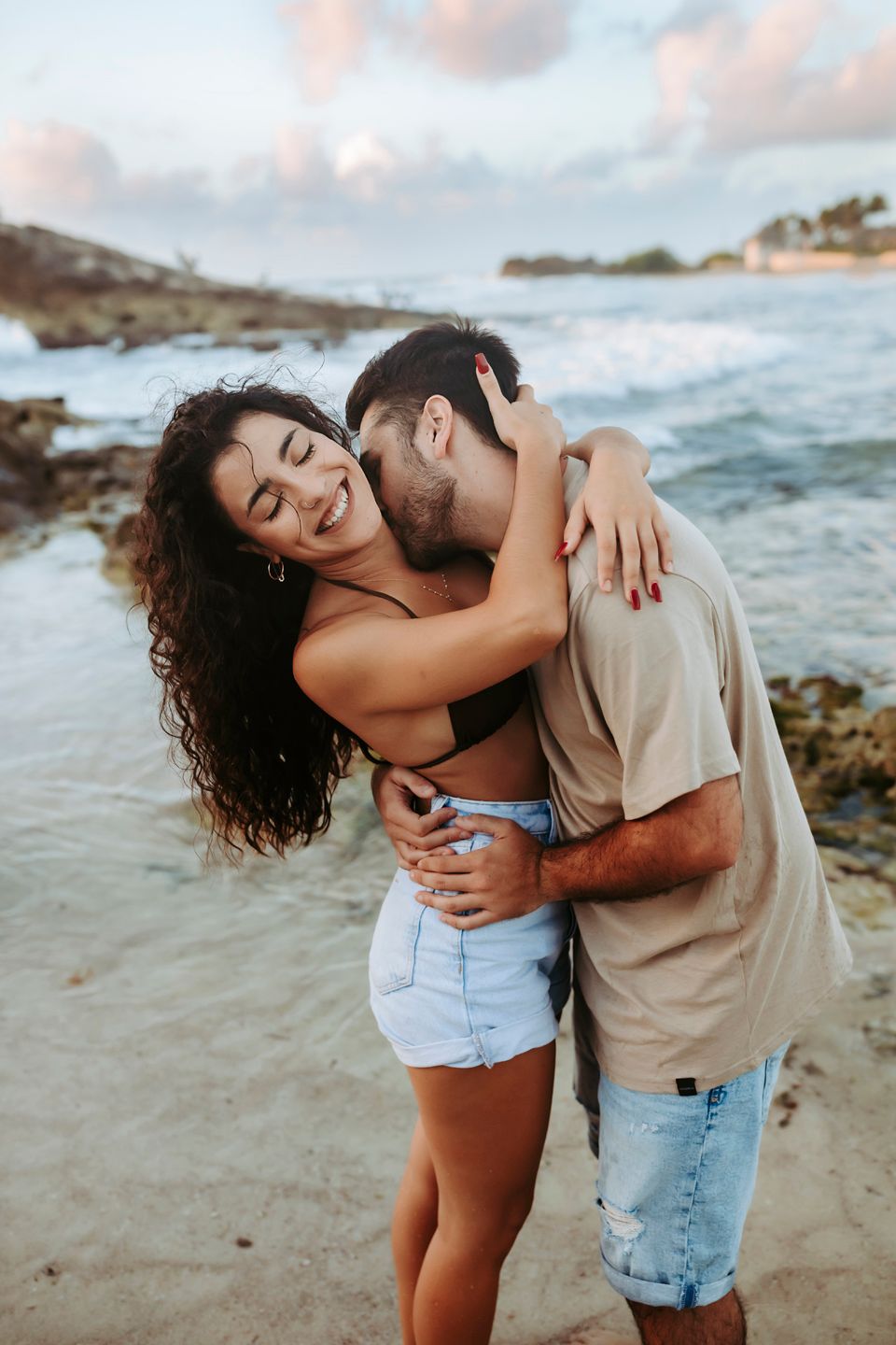 A romantic embrace between a couple on a beach with waves and rocks in the background.