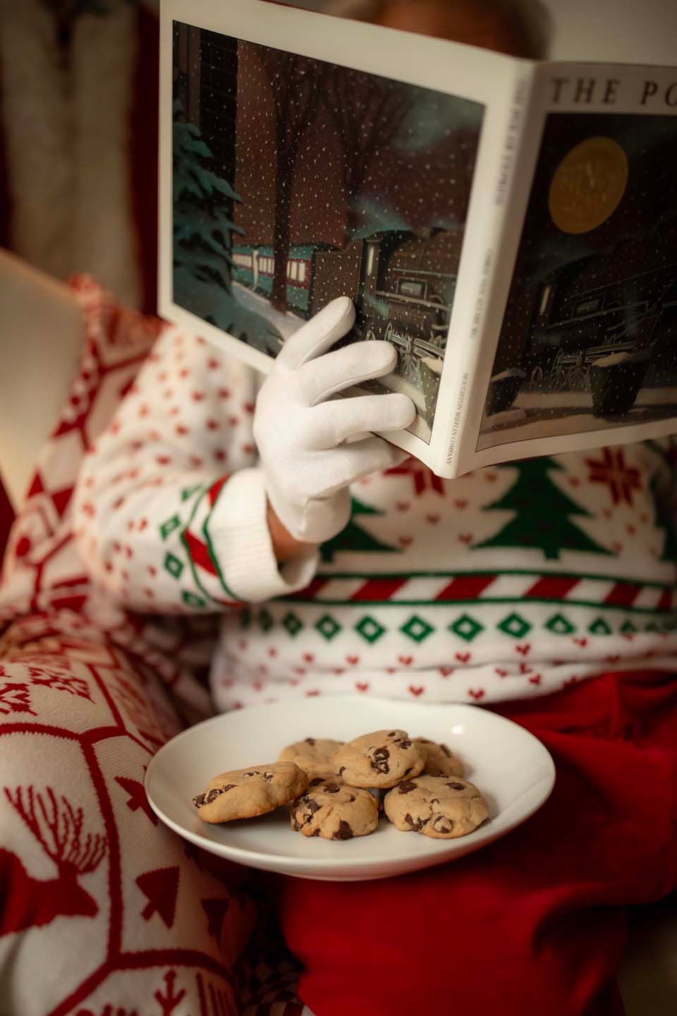 Santa Claus in Christmas sweater reads The Polar Express book beside a plate of chocolate chip cookies.