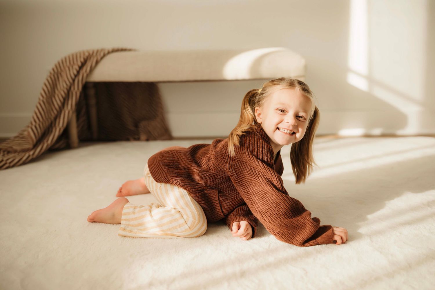 A child in a cozy brown sweater lies playfully on a sunlit white couch.