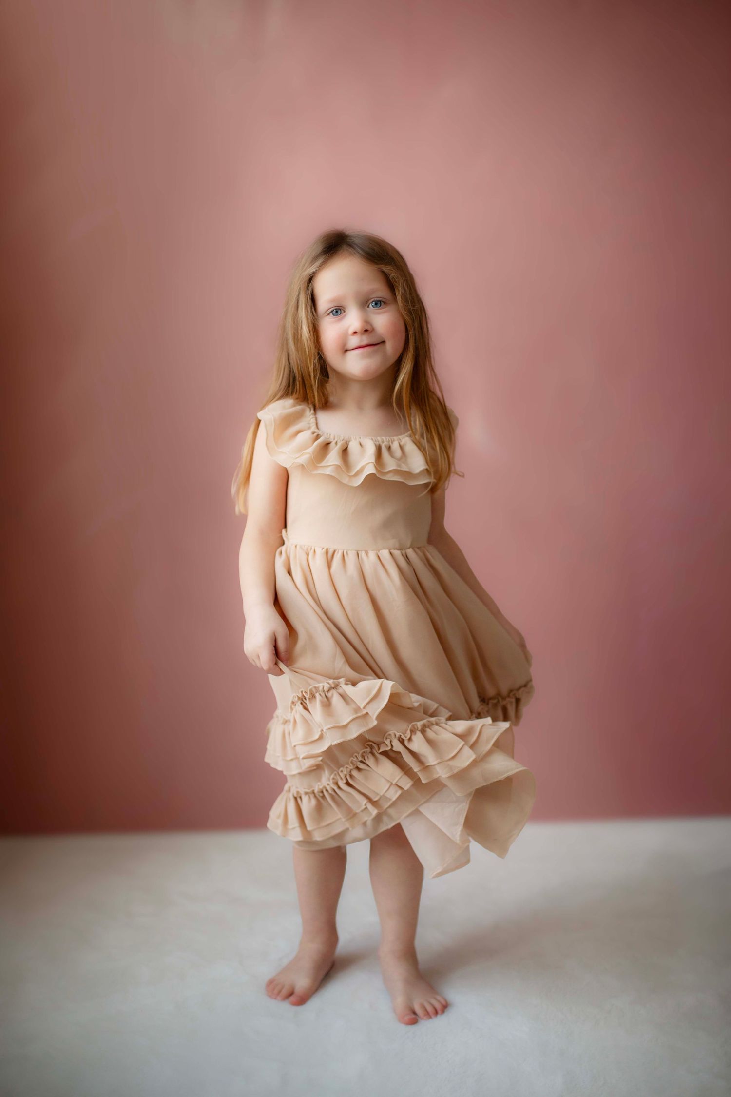 Young child wearing a flowing beige ruffle dress poses barefoot against a pink studio backdrop.