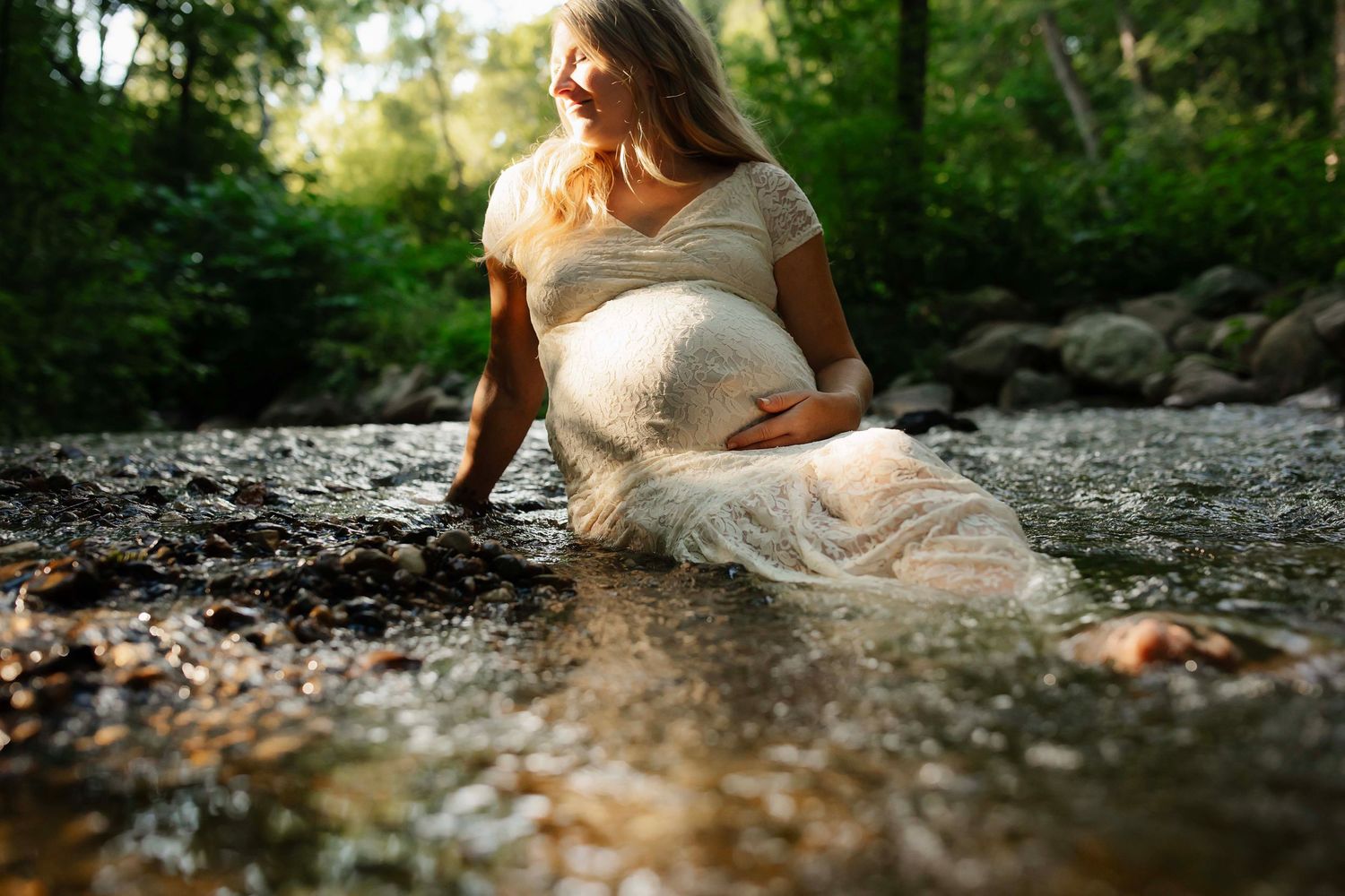 Pregnant woman in white dress sitting in a rocky stream surrounded by lush green trees during golden hour.