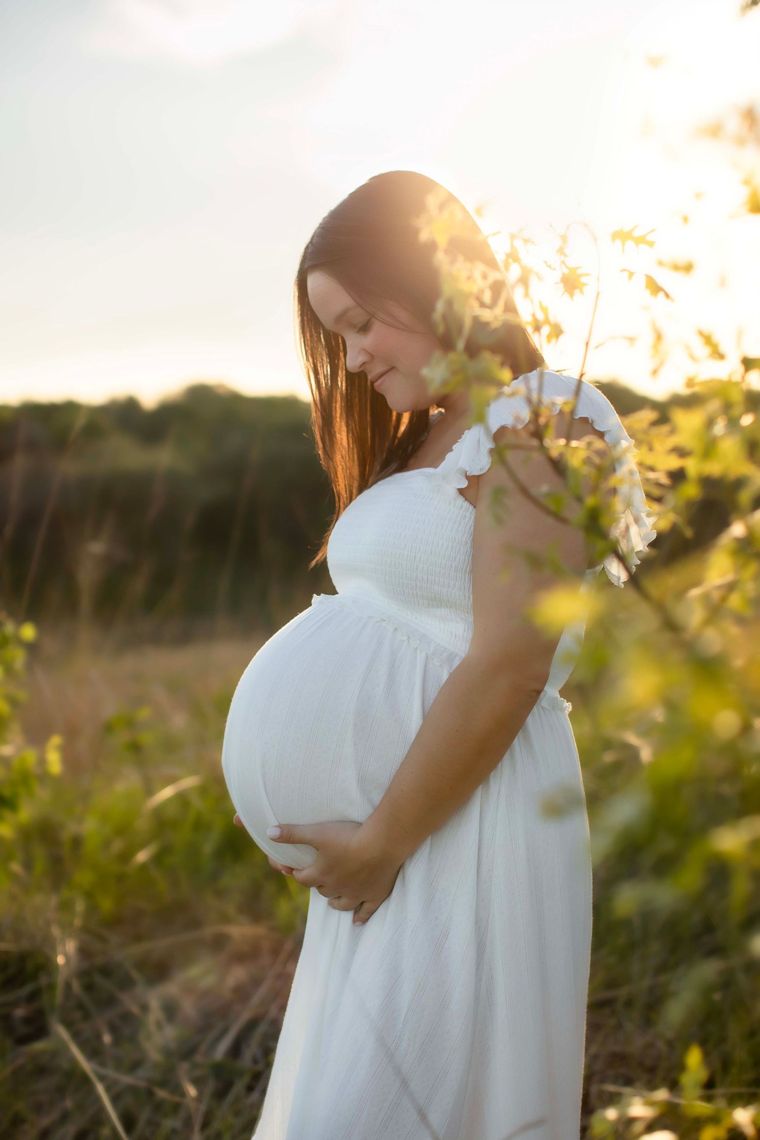 Pregnant woman in white dress holding baby bump outdoors in golden sunset light among wildflowers.