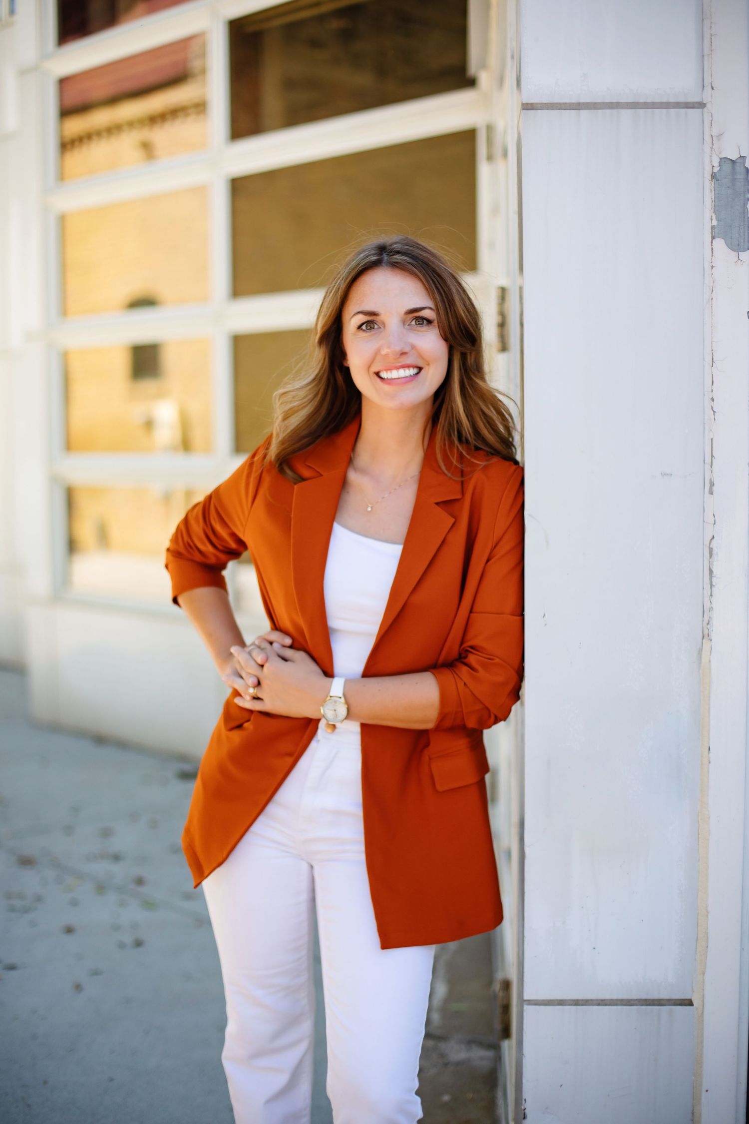 Stylish woman in orange blazer and white pants stands confidently against white wall.