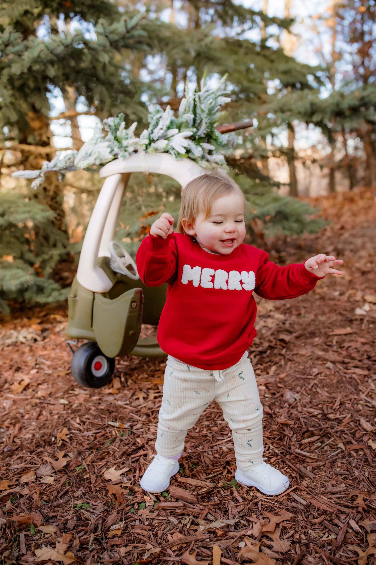 Toddler in red Merry sweater dancing by decorated cozy coupe car outdoors.