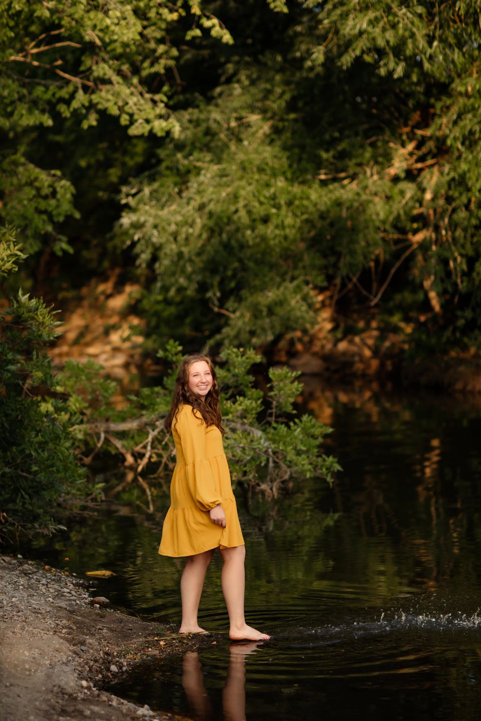 Person in a yellow dress standing barefoot in a creek surrounded by lush greenery on a sunny day.