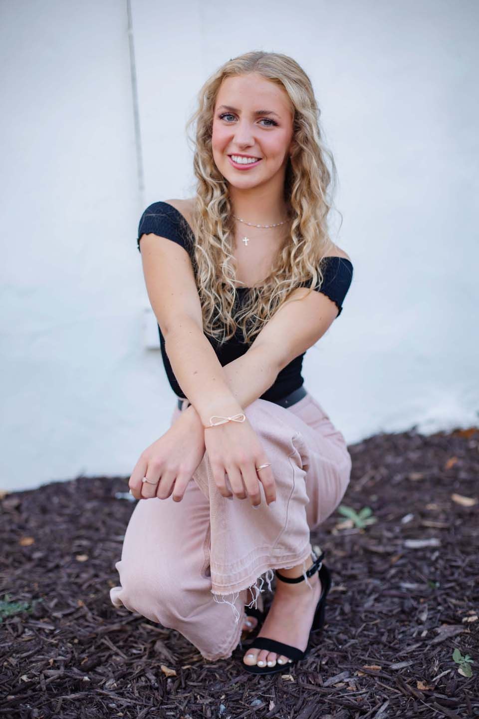 A casual portrait showing a black top and blush pants while crouching on mulch.