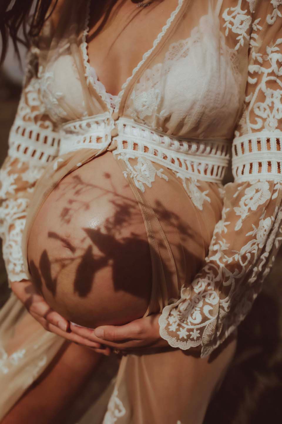 Close-up of pregnant belly being cradled in white lace dress with delicate patterns and floral embroidery details.