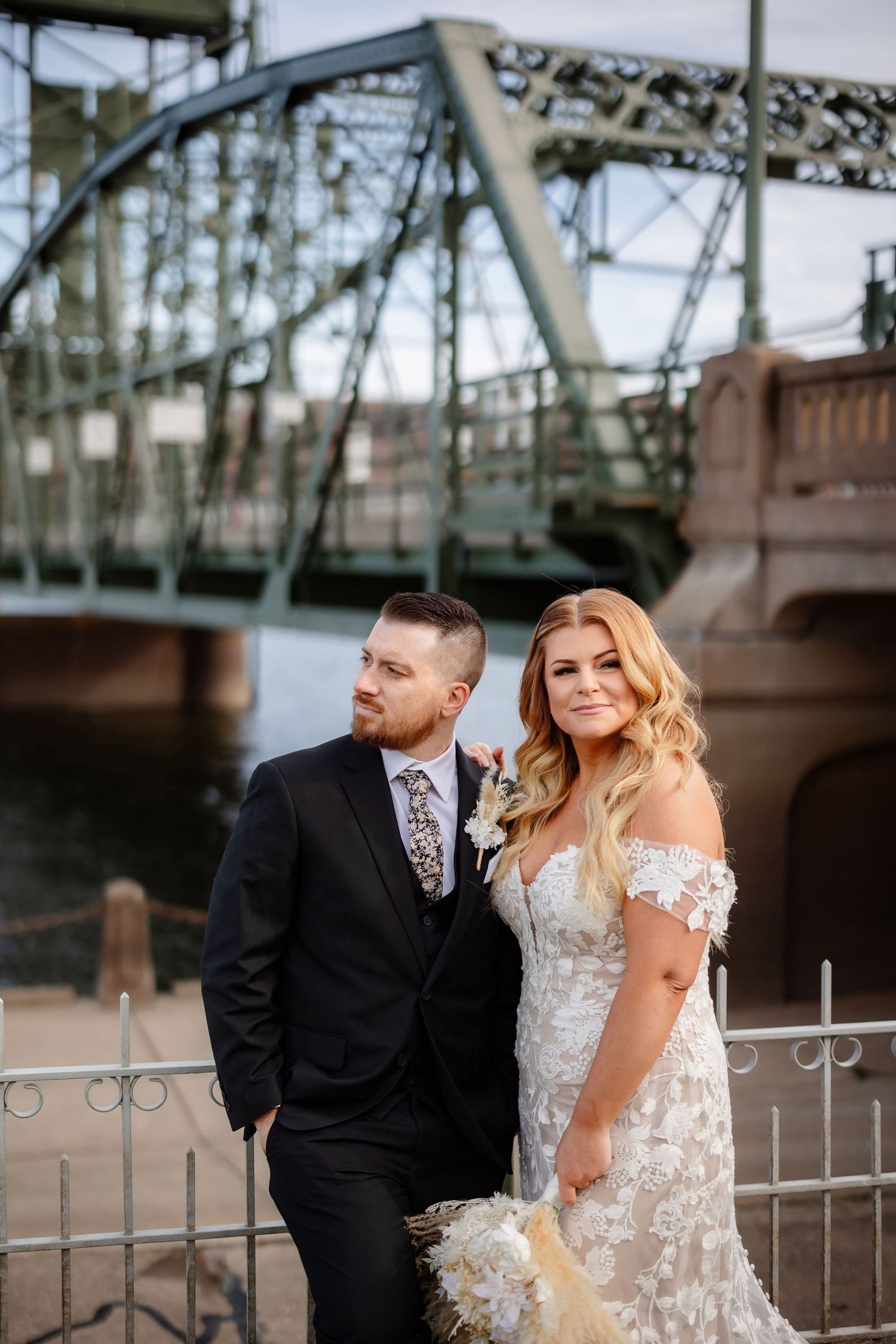 A couple poses for wedding photos in front of a historic steel bridge structure.