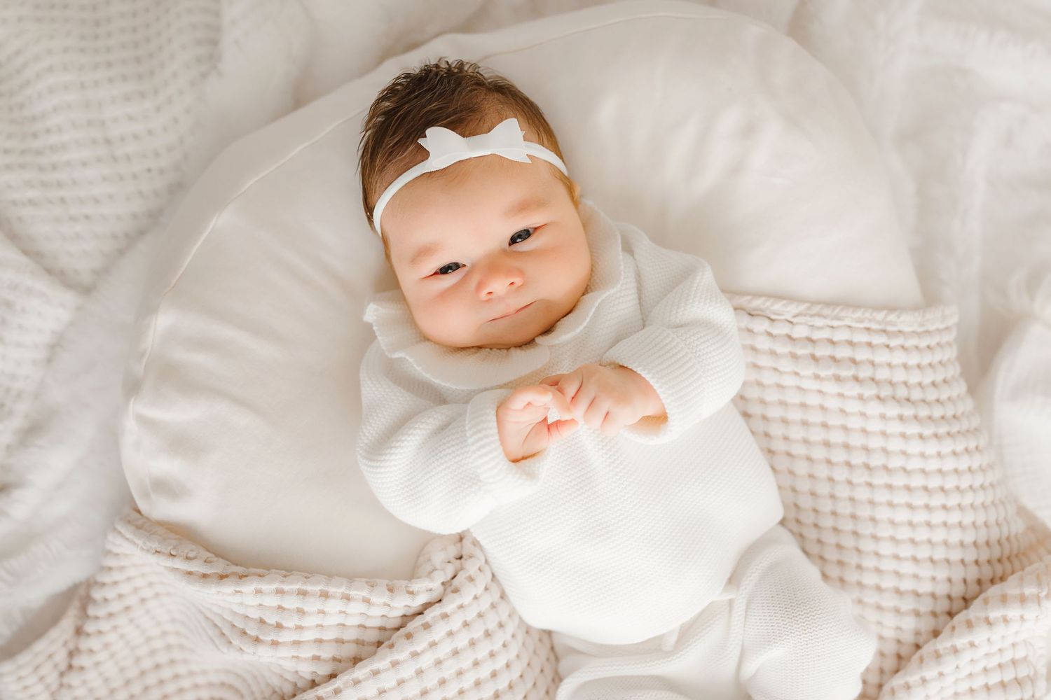 Baby in white outfit smiling while resting on textured white blanket.