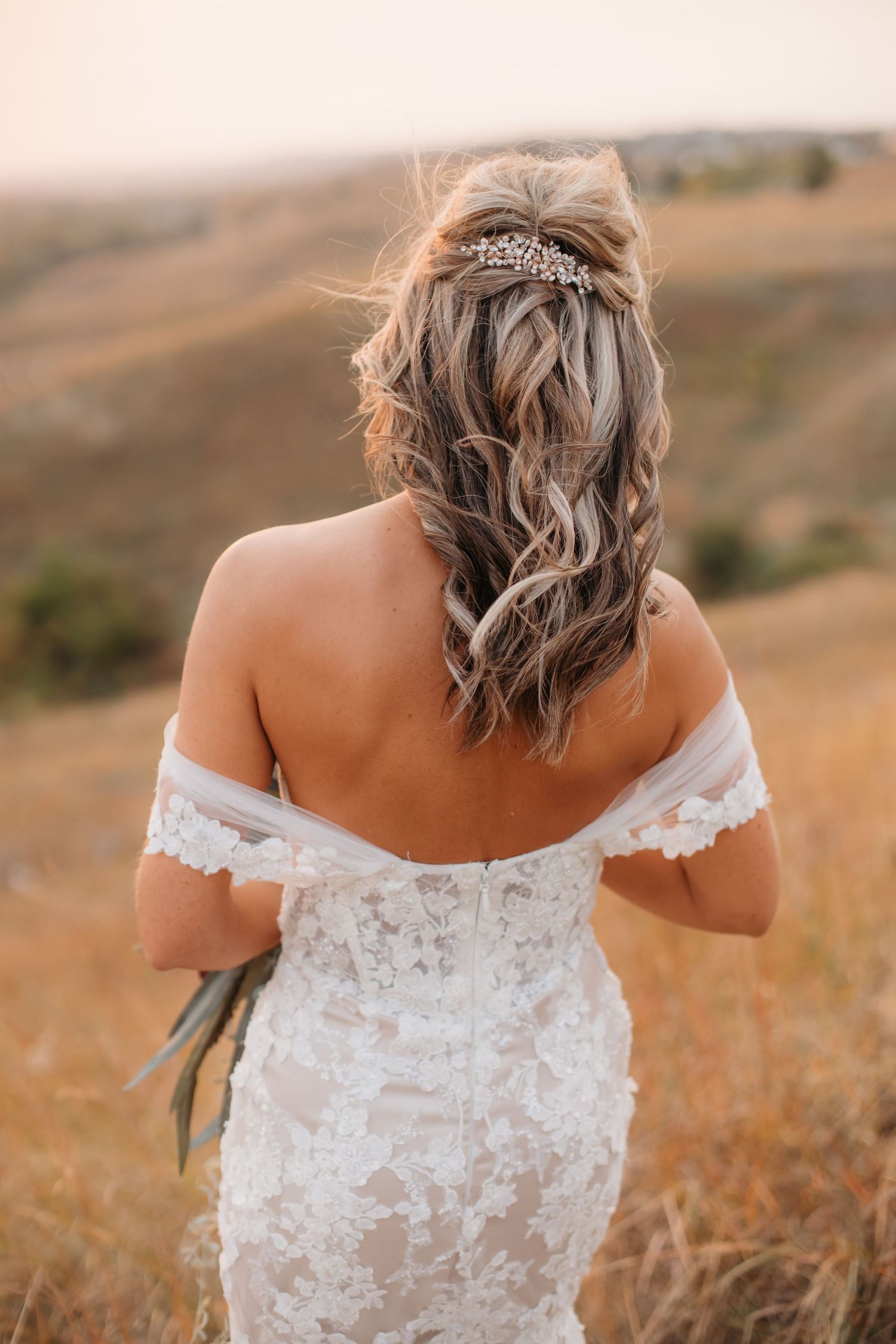 Back view of bride in lace off-shoulder wedding dress with crystal hair accessory in autumn field at sunset.